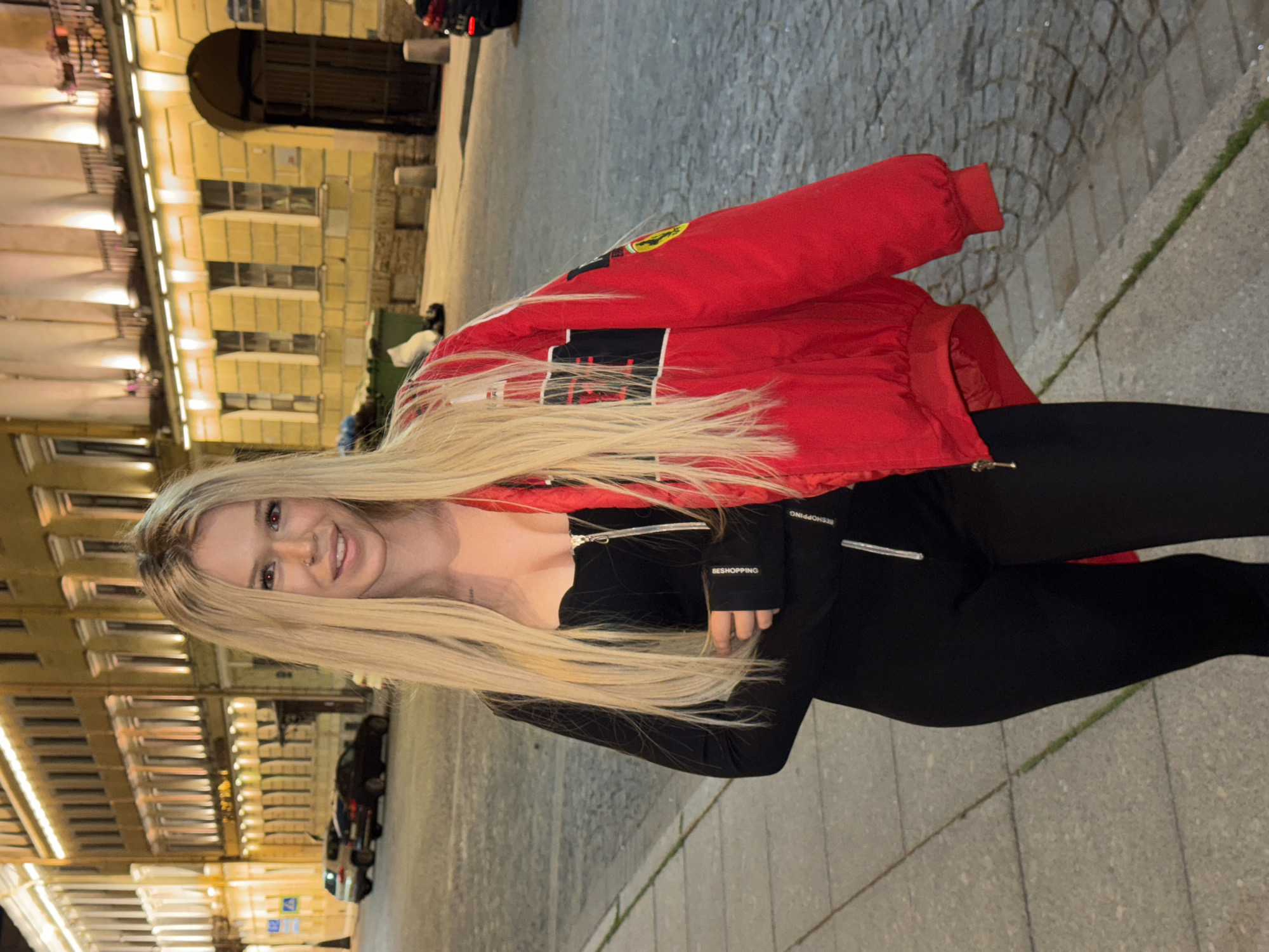 A young woman with long blonde hair stands outdoors at night in Saint Petersburg, Russia. She is wearing a red jacket over a black top and black pants. The jacket has "FERRARI" visible on the sleeve in red, yellow, and black. The black top has "BESHOPPING" written on it in white. She is smiling at the camera. Behind her, a cobblestone street is visible, and in the background, there are illuminated buildings with many windows, typical of Saint Petersburg's architecture. Cars are parked along the street, suggesting an urban environment. The lighting indicates it is evening or night.