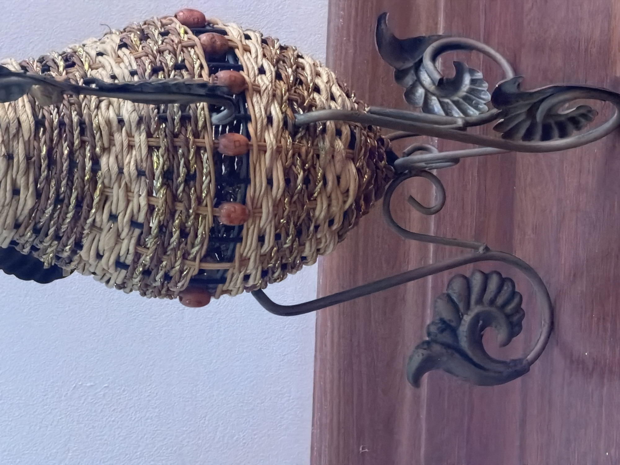 A woven basket, possibly made of straw and adorned with gold thread and small brown beads, sits on a metal stand with ornate, leaf-like feet. The basket has a dark rim at the top. The stand is dark grey or bronze. The background consists of a white textured wall and a wooden surface, likely a shelf or table, in the foreground. The lighting suggests an indoor setting. There are no people or animals visible in the image. The scene is stationary and depicts a decorative object. There is no information in the image to suggest it is located in Makhachkala, Russia.