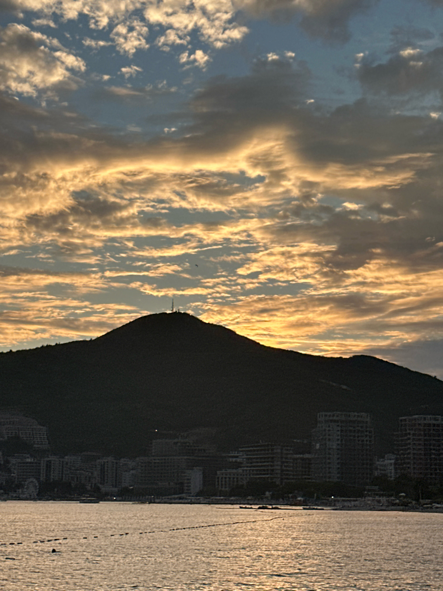 The image captures a coastal city scene at dusk, with a prominent silhouetted mountain overlooking the water and the cityscape. The sky is filled with dramatic clouds illuminated by the setting sun, casting a warm, golden glow. Several buildings, some appearing to be under construction, are visible along the coastline, nestled at the base of the mountain. The water in the foreground is calm, reflecting the light from the sky, and a series of buoys mark a section of the water. A lone antenna or tower is visible atop the mountain. While no people are clearly discernible, the presence of buoys suggests potential human activity in the water. The overall impression is one of a serene evening in a Mediterranean coastal town. Based on the distinctive silhouette of the mountain and the characteristic architecture, this scene is likely located in Budva, Montenegro, with the prominent peak possibly being the island of Sveti Nikola or a similar landmark visible from the mainland.