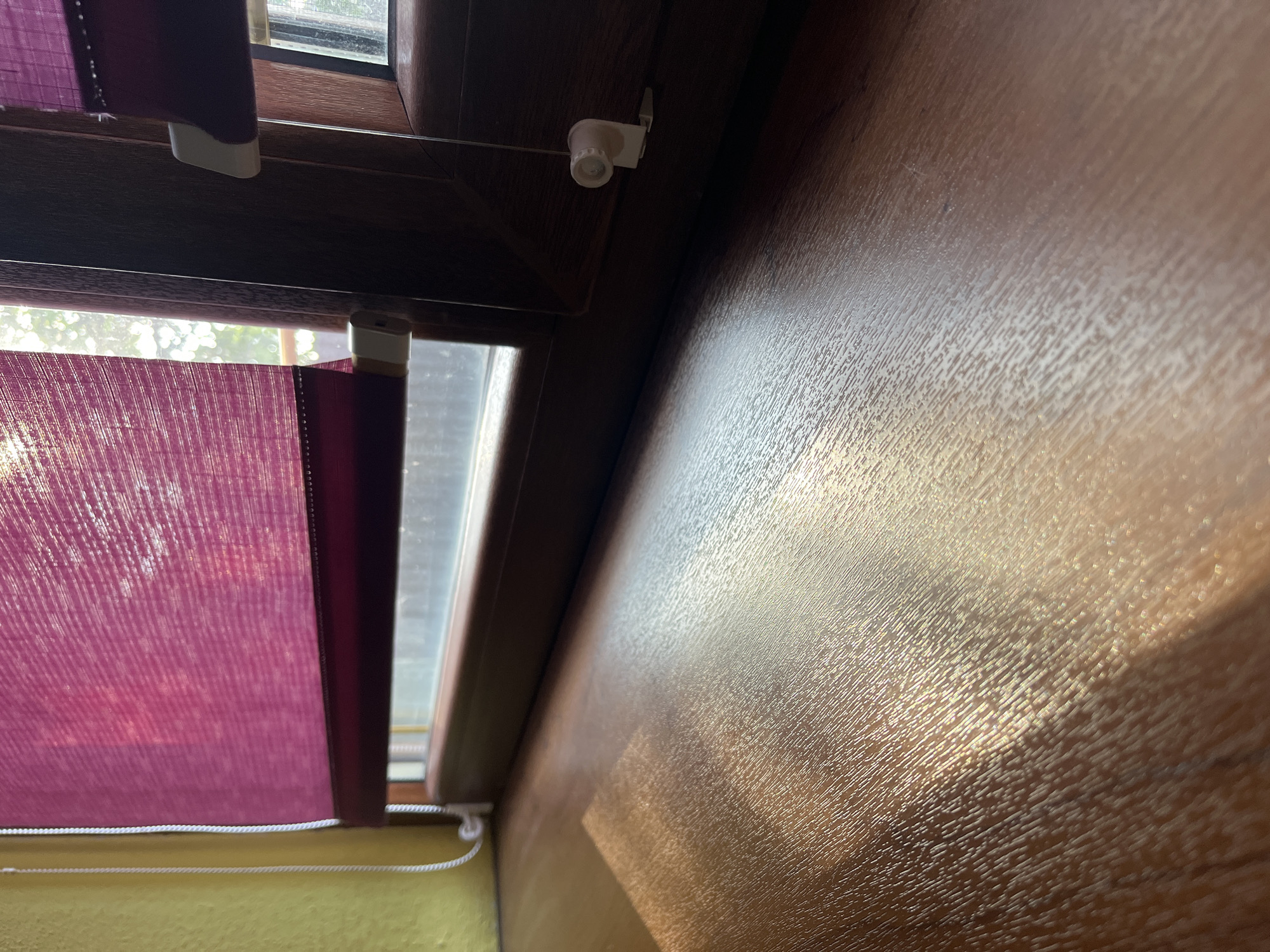 The image shows a close-up view of a wooden windowsill with sunlight reflecting on its surface. In the background, a window is visible with a purple roller blind partially lowered. The window frame is dark brown. The wall to the left of the window is painted yellow. There is no indication of the city of Tarnivtsi, Ukraine in the image, nor are there any people, actions, or interactions depicted.