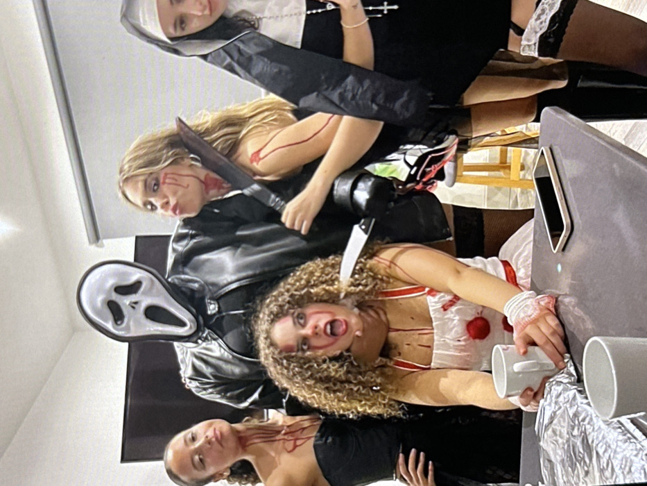 Within an indoor setting in Novosergievka, Russia, five individuals are arranged, posing for a photograph in Halloween costumes. In the central background stands a person dressed as Ghostface, wearing a black hooded robe and a black leather jacket, with the characteristic white Scream mask covering their face. To the left of Ghostface, a woman with dark hair and skin, wearing a black strapless top, has artificial blood streaking her neck and chest. She poses with her right hand on her hip, looking forward.
In the immediate foreground, centered, a woman with curly brown hair and fair skin wears a white, blood-splattered costume with red accents, resembling a clown or nurse. A fake knife is positioned at her throat, seemingly held by the Ghostface figure, and her mouth is open in an expression of distress. Her bandaged wrists, also blood-stained, rest on a dark grey table, where she holds a white mug. To the right of Ghostface, a woman with long blonde hair and fair skin wears a dark top, with artificial blood on her cheek and neck. She holds a large, fake cleaver-style knife in her right hand and pouts towards the camera with her left hand raised. Further to the right, a woman in a black and white nun habit and robe, holding a rosary, also has artificial blood visible on her face. Both the blonde woman and the nun are visible wearing dark thigh-high stockings, with the nun's featuring white lace tops.
The dark grey table in the foreground also holds a black mobile phone and an object wrapped in silver foil. The background includes a dark flat-screen television screen, a white wall, and a window fitted with white horizontal blinds. A light-colored wooden stool is partially visible on the right.