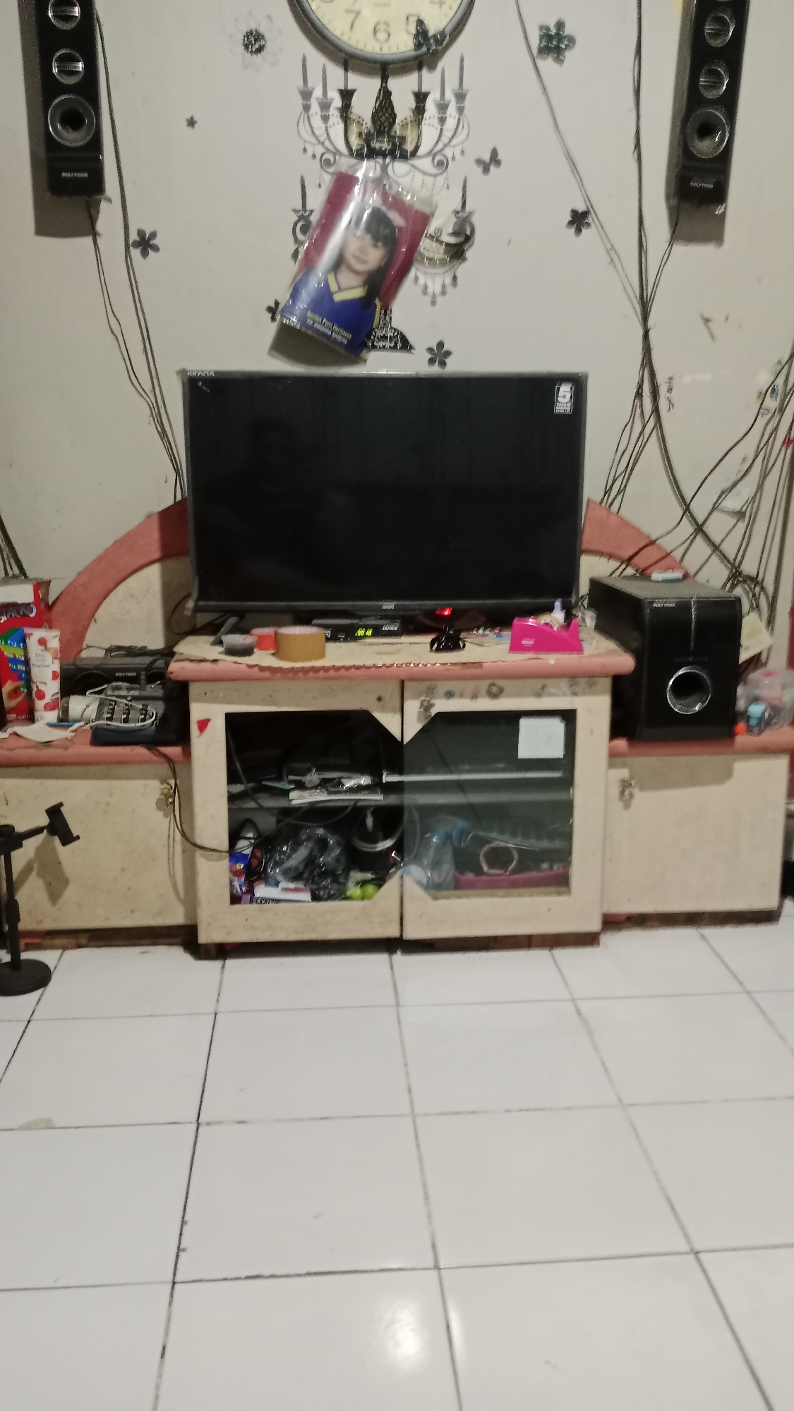The image depicts an indoor scene, likely a living room or entertainment area, set in the city of Blitar, Indonesia. Prominently featured is a large black television set resting on a wooden entertainment stand. The stand itself has several compartments, some of which are visible and contain various items, including what appears to be electronic devices and possibly some bagged goods. To the left of the television, a dark-colored speaker is mounted on the wall, and another similar speaker is on the right side of the entertainment stand. Above the television, a framed picture of a young girl is displayed, leaning against the wall. The wall itself is decorated with what appears to be wallpaper or decals, including images of a chandelier and floral motifs. A large analog clock with Roman numerals is also visible on the wall. The floor is tiled with white square tiles. The overall impression is of a lived-in space with personal belongings present.