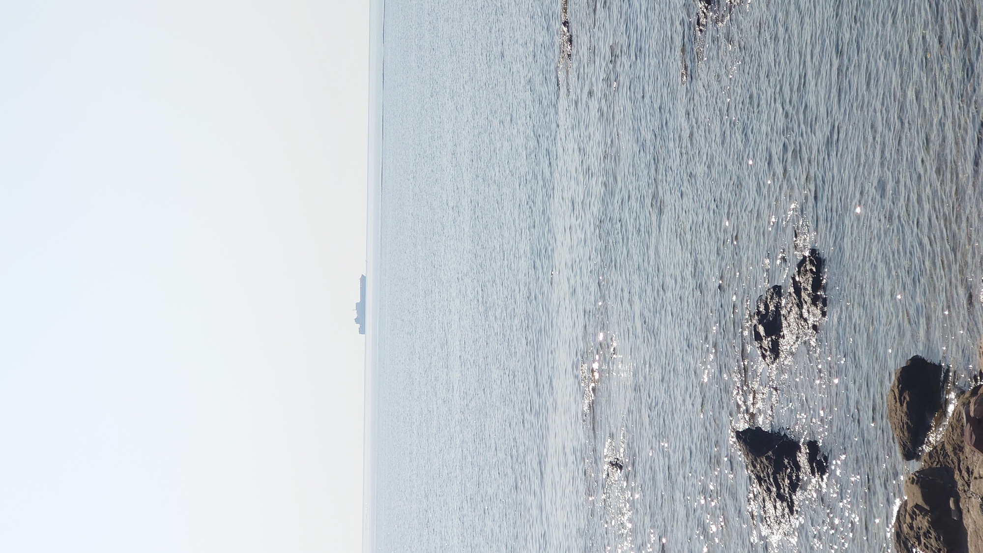 The image depicts a serene coastal scene with a vast expanse of calm sea under a clear, bright sky. In the foreground, dark, wet rocks are partially submerged in the shallow water, with sunlight glinting off the surface. Further out in the middle ground, a large cargo ship is visible on the horizon, silhouetted against the bright sky. The water in the midground and background is a soft, muted blue, reflecting the clear sky. The overall impression is one of tranquility and vastness. The location is the coastline of Biddeputzi/Villaputzu, Italy.
