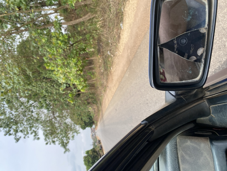 A view from inside a moving vehicle in Magongo, Nigeria, shows a two-lane paved road extending into the distance under an overcast sky. The vehicle's dark-colored dashboard and a-pillar are visible in the lower left. A black-framed side mirror on the right exhibits significant damage, with a large, roughly triangular section of its glass missing from the upper center, and the remaining surface displaying multiple circular depressions and minor glass fragments.
On the immediate right of the road, a dirt shoulder is present, beyond which a dense line of various trees and green foliage rises, some appearing deciduous. Several thin, vertical posts are interspersed within this vegetation. Debris, including white fragments, is scattered on the ground alongside the shoulder. On the distant left side of the road, several dark, indistinct bovine figures are visible, along with more trees and structures. The road itself appears somewhat aged and extends toward a gentle curve to the left, disappearing over a slight rise in the far distance. No people are explicitly visible.