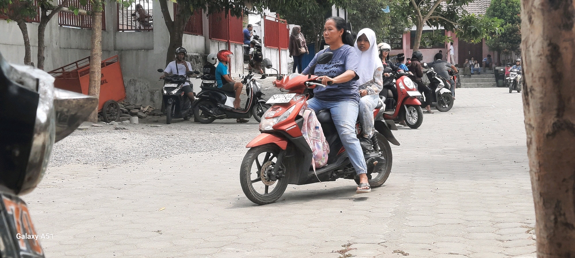 In Tangkil, Indonesia, a woman and a young girl ride on a red scooter, the woman driving and the girl seated behind her. The woman is wearing a blue t-shirt and jeans, and the girl is wearing a white hijab and a patterned shirt. They are traveling on a paved road with a sidewalk and some trees in the background. Several other motorcycles and people are visible in the scene. An orange bin is on the left side of the image.