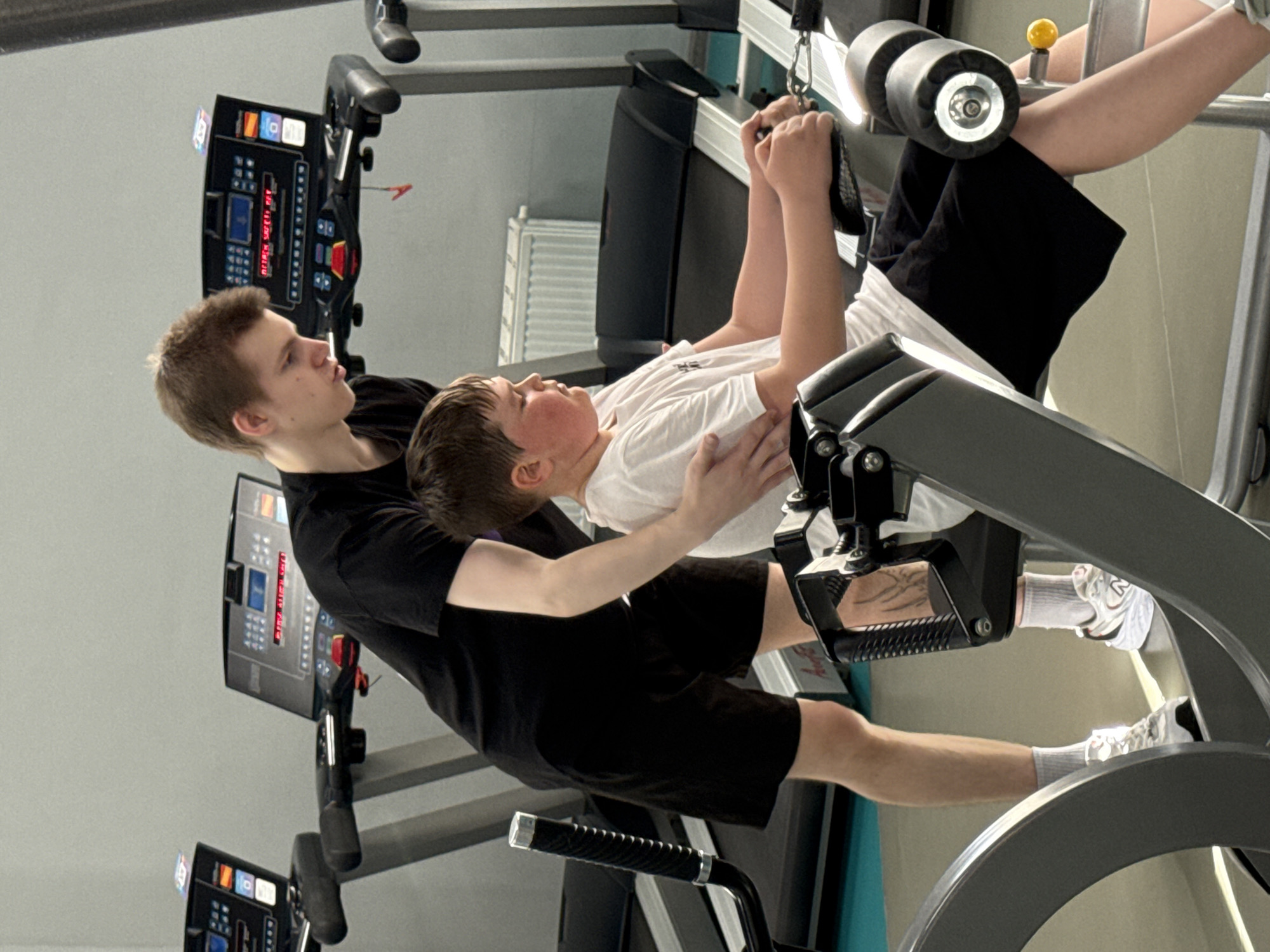 In an indoor gym in Yekaterinburg, Russia, two males are engaged in a fitness activity. A younger male, appearing to be a child or pre-teen, is lying on his back on a leg press or hip sled machine, pushing the footplate upwards with his legs. He is wearing a white t-shirt, black shorts, white socks, and white athletic shoes, which have "Hoka" visible on one. His hands are gripping the machine's handles above his head.
An older male, likely a teenager or young adult, stands behind and to the side of the younger boy, spotting him. He has his hands placed on the younger boy's lower back or waist, providing support and guidance during the exercise. The older male is dressed in a black t-shirt and black shorts. Both individuals display focused expressions, concentrating on the movement.
In the background, several pieces of gym equipment are visible, including at least two treadmill consoles. These consoles feature various buttons and digital displays showing fitness metrics such as "SPEED," "DISTANCE," "CALORIES," "TIME," "PULSE," and "INCLINE." A red digital readout on one console displays numerical values, and the text "SAFETY KEY" is partially discernible. The overall setting is brightly lit, typical of a fitness center.
