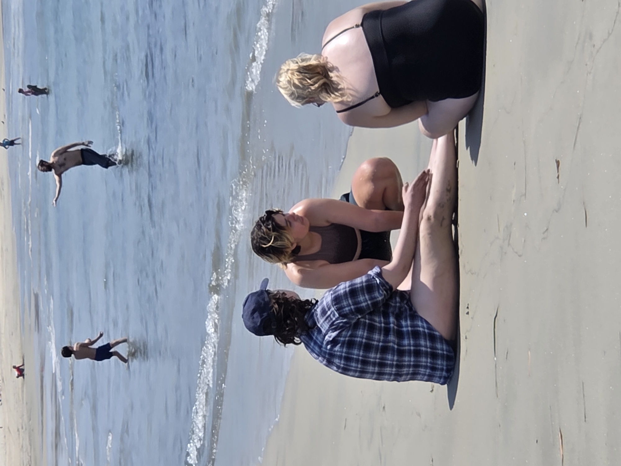 The image depicts a sunny day at the beach in Long Beach, United States of America. In the foreground, three individuals are seated on the sand. The person on the left, wearing a blue and white plaid shirt and a blue baseball cap, is facing away from the camera and toward the ocean. In the middle, another person is crouched, facing partially towards the person on the left, wearing a dark-colored top. To the right, a third person, seen from behind, is wearing a black swimsuit. The ocean is visible in the background with gentle waves breaking on the shore. Further out in the water, several other people are visible. One individual, a child in dark shorts, stands in the shallow water. Another person, an adult male with a beard, is wading in the water, extending an arm. Several other figures are also present in the water at a distance. The beach appears sandy and clean, with the ocean water being a clear blue.