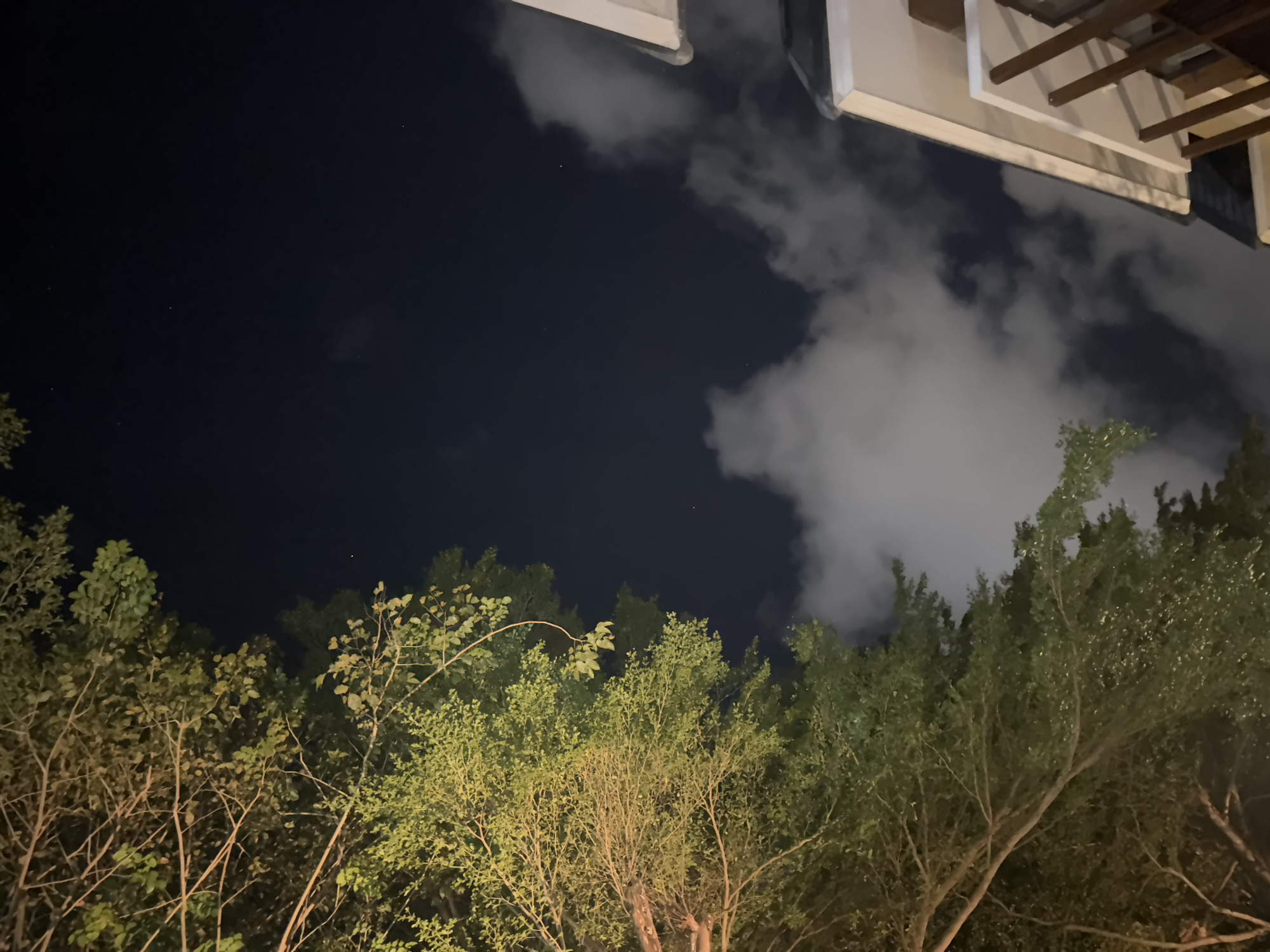 The image presents a low-angle view, primarily capturing the night sky and the tops of trees, likely in Kuta Selatan, Indonesia. The sky is a mix of dark blue and cloudy patches. A portion of a building, possibly a residence or hotel, is visible in the upper right corner, featuring light-colored walls and a wooden pergola. Below the sky, a dense cluster of trees fills the lower portion of the image. The trees appear to be various species, their leaves reflecting light, possibly from artificial sources. No people or direct actions are visible in the frame. The environment suggests a residential or resort area in a tropical setting.