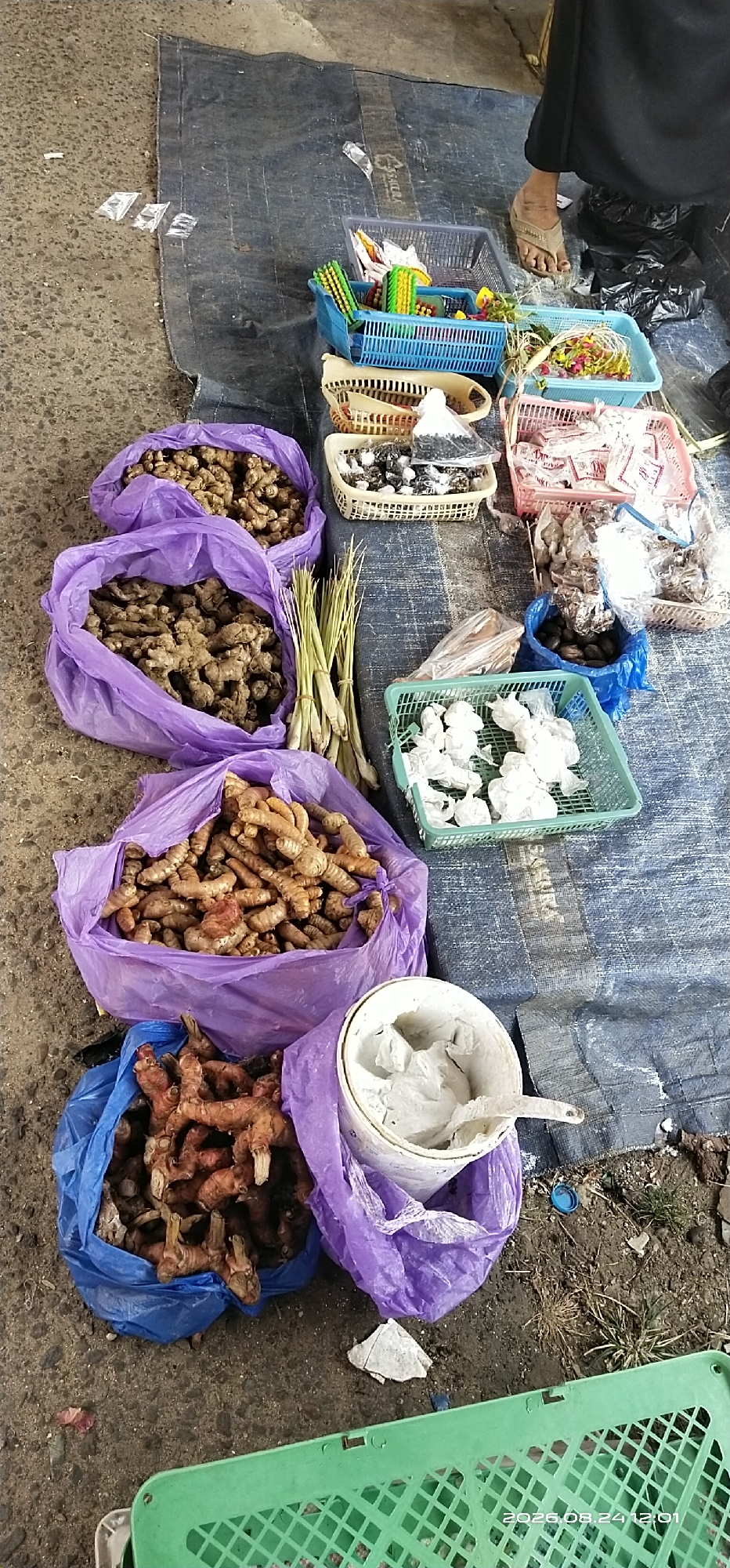This is a high-angle, vertical shot of a street market stall in Tamiang Layang, Indonesia. The ground is covered by a dark blue tarp, upon which various goods are displayed. In the foreground, there are several large purple plastic bags filled with fresh ginger and galangal roots. To the right of these bags is a light blue plastic bag also containing galangal. Next to the purple bags, a bunch of lemongrass is laid out. Further to the right, a white plastic bucket with a scoop contains a white paste or powder.
Scattered around the tarp are several plastic baskets of various colors, containing different items. Some baskets hold what appear to be packaged goods, possibly spices or condiments, while others contain darker, granular items and some white powdery substances in plastic bags. A person's foot, wearing a brown sandal, is visible in the upper right portion of the image, suggesting the stall is actively being managed. The overall scene depicts a typical outdoor market setting.