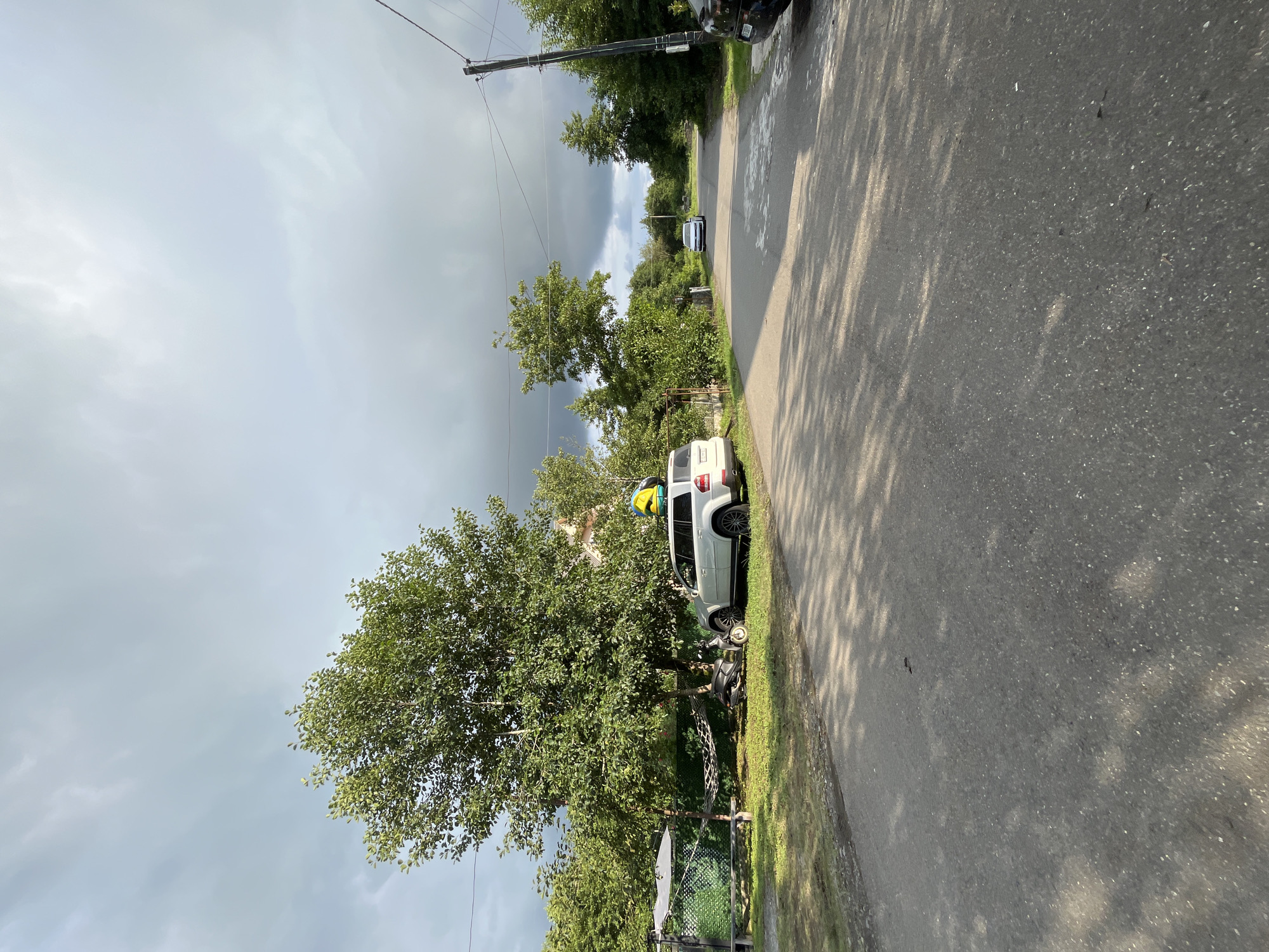 A white SUV is parked on the side of a paved road in Ureki, Georgia. The SUV has a yellow and blue object on its roof. A black scooter is parked next to the SUV. A hammock is suspended between two poles near the vehicles. The road is lined with green trees and vegetation. The sky is cloudy. In the distance, a white car is driving down the road, and a power pole with wires is visible on the right side of the road.