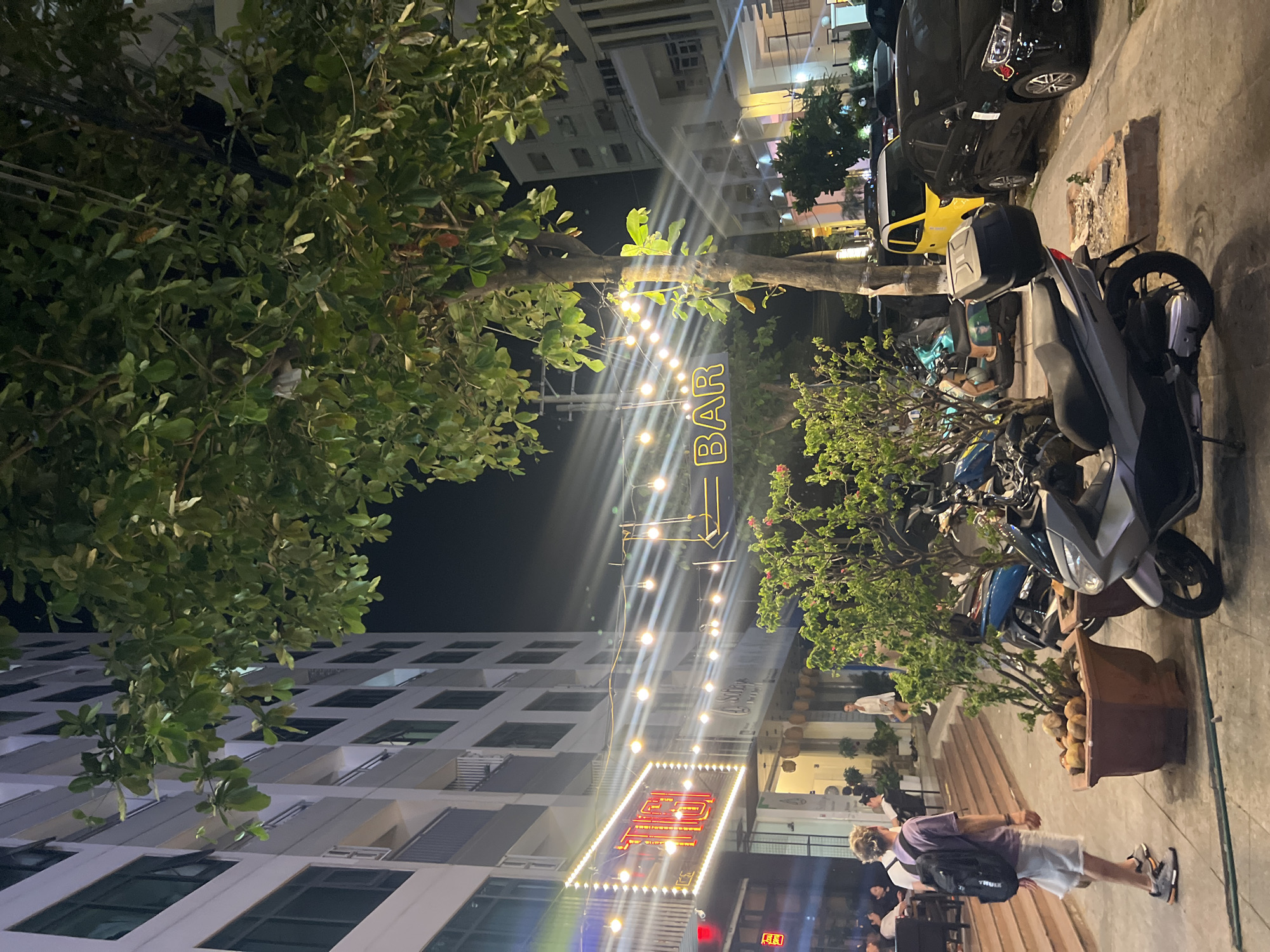The image depicts an outdoor scene at night in what appears to be Bắc Nha Trang, Vietnam. The perspective is from street level, tilted upwards.
Buildings: A tall, white building with numerous windows is visible on the left, while a similar building is seen in the background, closer to the center.
Signage: Two illuminated signs are prominent. One reads "BAR" vertically with string lights emanating from it. The second, closer to the bottom of the frame, has Vietnamese script, also lit up.
Street Scene: Several parked motorbikes are visible on the right side of the image. Pedestrians are walking on the sidewalk.
Vegetation: Trees with lush green foliage partially obscure the view on the left and right sides of the frame. One tree appears to have string lights attached to it near the "BAR" sign.
Lighting: The scene is lit by streetlights, the illuminated signs, and string lights.
People: At least one person can be seen from the waist up, walking along the sidewalk. Additional people are partially visible in the lower portion of the frame.
