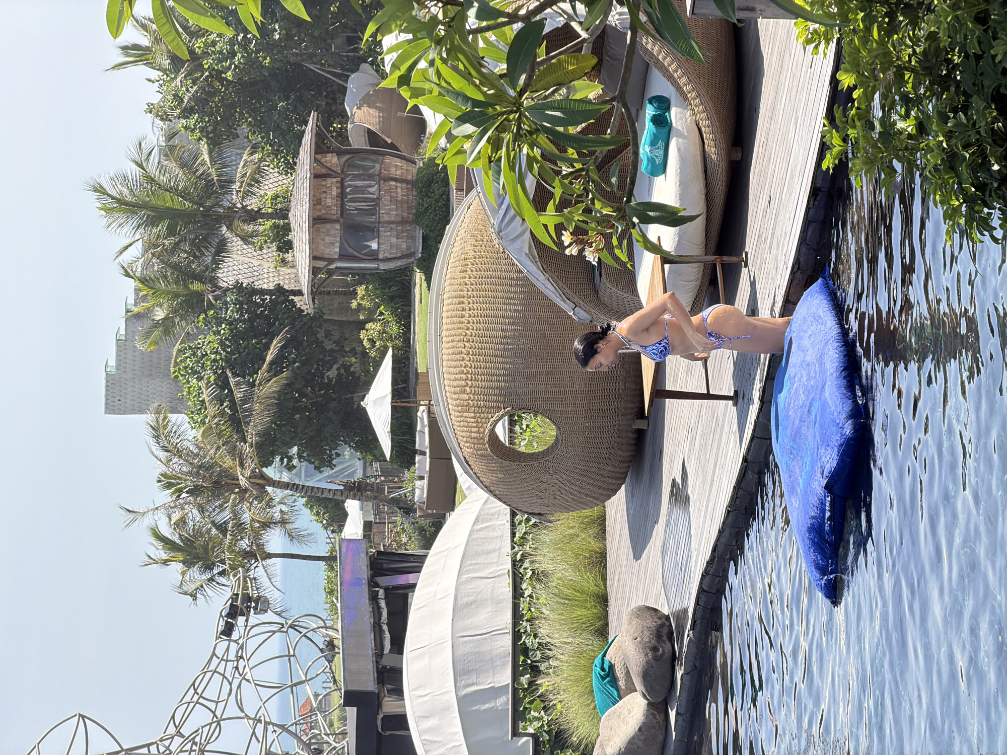 A woman in a blue and white bikini stands at the edge of a swimming pool. She is looking down and to her right, with her hands on her hips. The pool is surrounded by a wooden deck. On the deck, there are several large rocks, a blue inflatable raft, and a table with a wooden top and metal legs. In the background, there are palm trees, a large woven structure, and a small thatched-roof building. A white umbrella is visible behind the building. The overall setting appears to be a resort or a private residence.