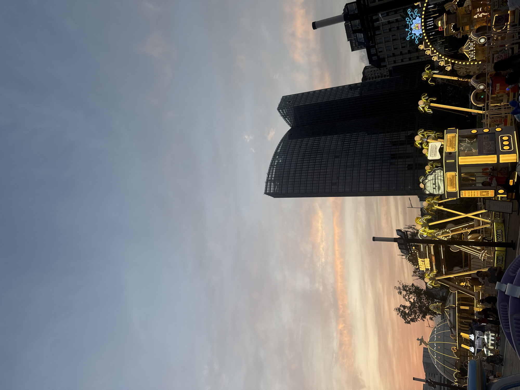 The image depicts an outdoor amusement park at dusk, with several rides illuminated by yellow lights. In the background, a modern, dark glass skyscraper with a curved facade dominates the skyline. The sky displays a gradient of blues and soft pinks, indicative of sunset. Several amusement rides are visible, including a swing ride with a sign that reads "HAPPY SWING" and a carousel-like structure with illuminated palm trees. People are present, some appearing to be enjoying the rides or waiting in line. The overall scene suggests a lively evening at an amusement park located in Shenyang City, China, with the prominent skyscraper serving as a landmark.