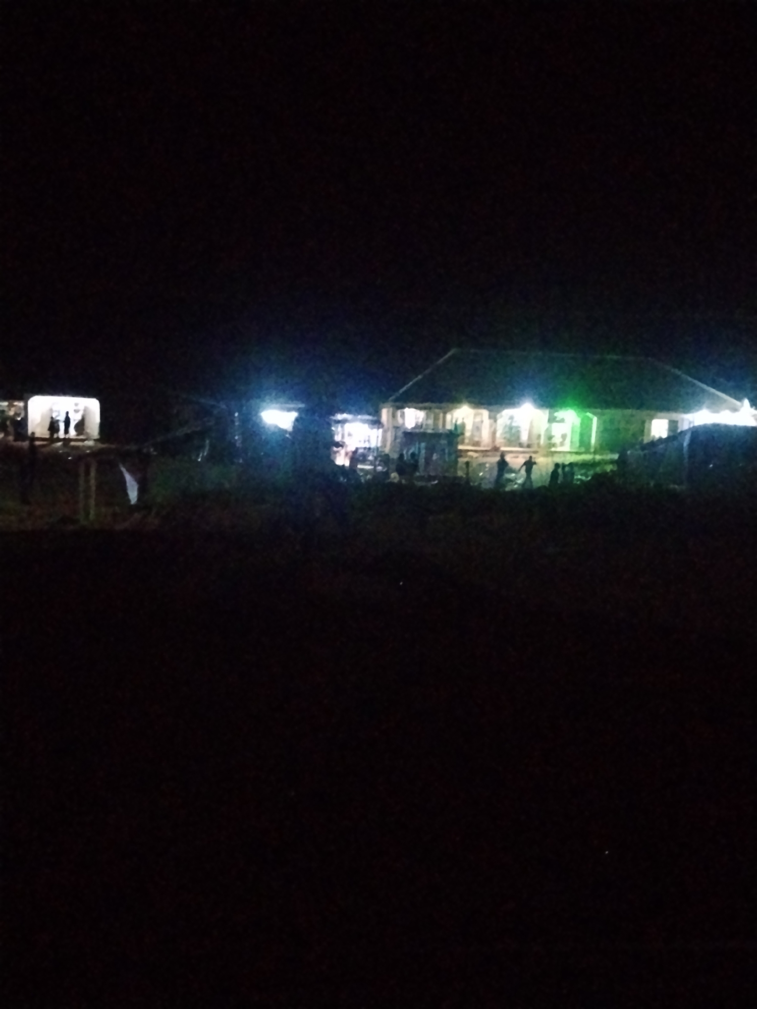 The image, taken at night in Durumi, Nigeria, shows a partially lit building with people present. The building appears to be a single-story structure with a pitched roof, illuminated by several bright lights, some of which cast a greenish hue. Silhouettes of people are visible in front of and inside the building, suggesting activity or gathering. To the left, another smaller structure or tent is also illuminated, with a few figures standing outside it. The foreground is dark and indistinct, likely representing unlit ground with some vegetation. The overall impression is of a public or community space that is operational after dark.