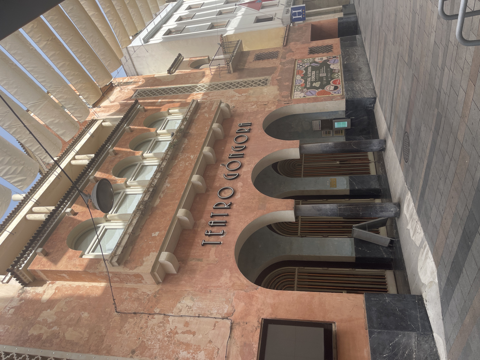 The image shows the exterior of the Teatro Góngora in Córdoba, Spain. The building has a weathered, peachy-pink facade with large arched doorways at street level, framed by dark marble. Above the arches are a series of windows and decorative architectural elements. A sign with the name "TEATRO GÓNGORA" is prominently displayed on the facade. A colorful poster for the "FESTIVAL GUITARRA CÓRDOBA" is visible on the right side of the building. A shaded walkway with patterned paving stones runs in front of the theater. The sky is visible above, with a series of white awnings stretched across the street. No people are visible in the image.