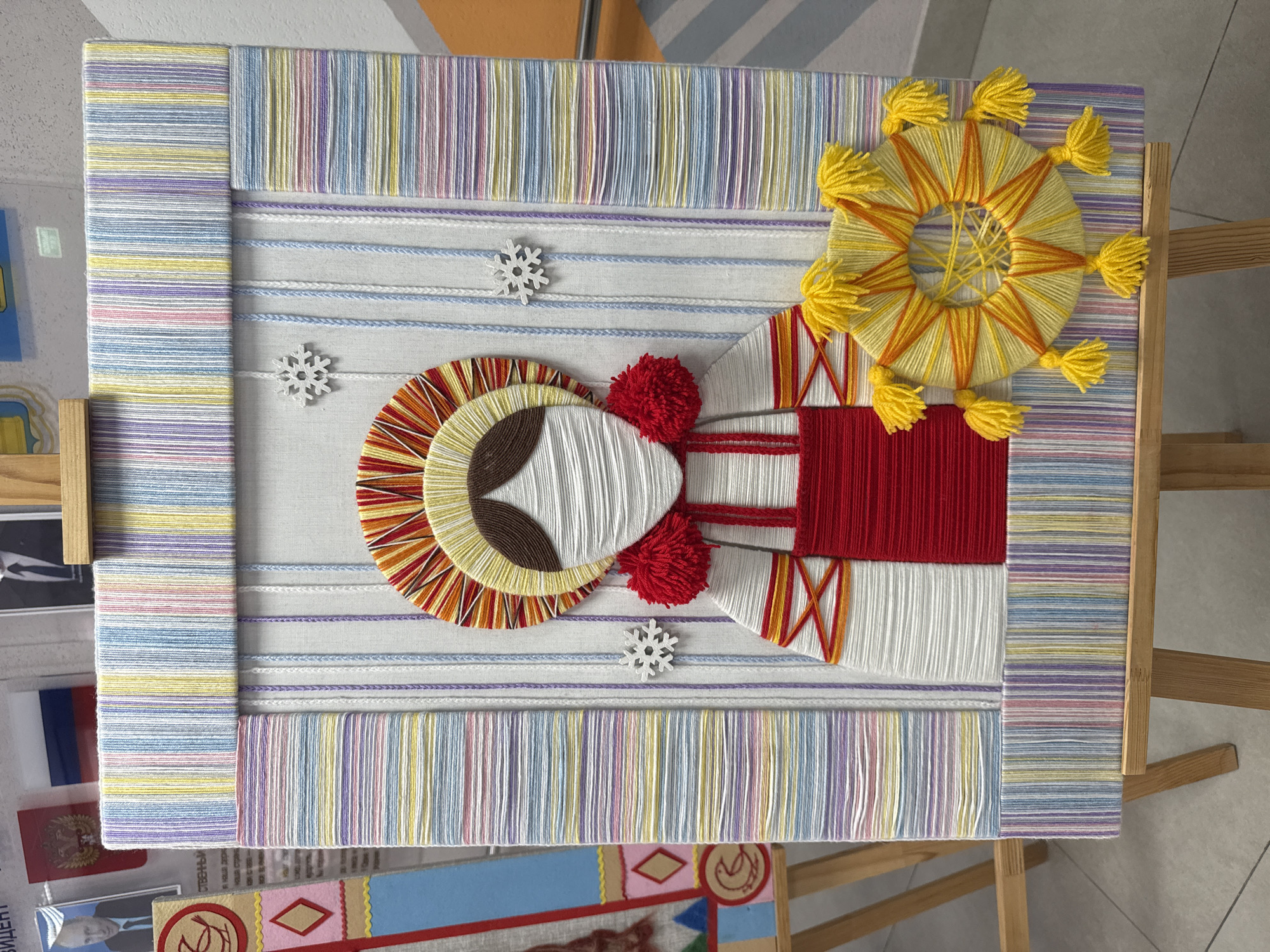 A framed craft project depicting a person with a sun is displayed on a wooden easel. The project is constructed from yarn and thread. The frame surrounding the project is made of yarn with stripes of color. The person has a white yarn face and body, brown yarn hair, and a headdress made of yellow and orange yarn. Red yarn pom-poms are attached to the headdress. The person wears a red garment with orange yarn accents. A large sun made of yellow and orange yarn with tassels is positioned above and to the right of the person. Three white snowflakes are attached to the background. The project is likely located in Tobolsk, Russia due to the flags and pictures of President Putin visible in the background.