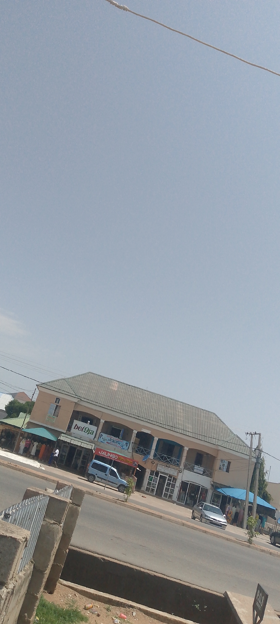 The image shows a street scene in Jalingo, Nigeria. In the foreground, a concrete barrier with metal railings runs along the side of a road. The road itself is paved and appears to be a main thoroughfare. On the right side of the road, there is a ditch or drainage channel.
Across the street, a two-story building with a light-colored facade and a grey, sloped roof dominates the middle ground. The building has several businesses on the ground floor, indicated by signs. One prominent sign reads "bet9ja". Another sign says "JALINGO Wine Store". There are awnings and entrances visible, with some people standing under them. The upper floor of the building features a balcony with railings.
Several vehicles are present on the street, including a light blue van and a silver sedan. People are seen walking on the sidewalk in front of the businesses. The environment appears to be a commercial area in a city. The sky is clear and light blue, suggesting a sunny day. The image is taken from a slightly elevated perspective, looking down at the street and building.