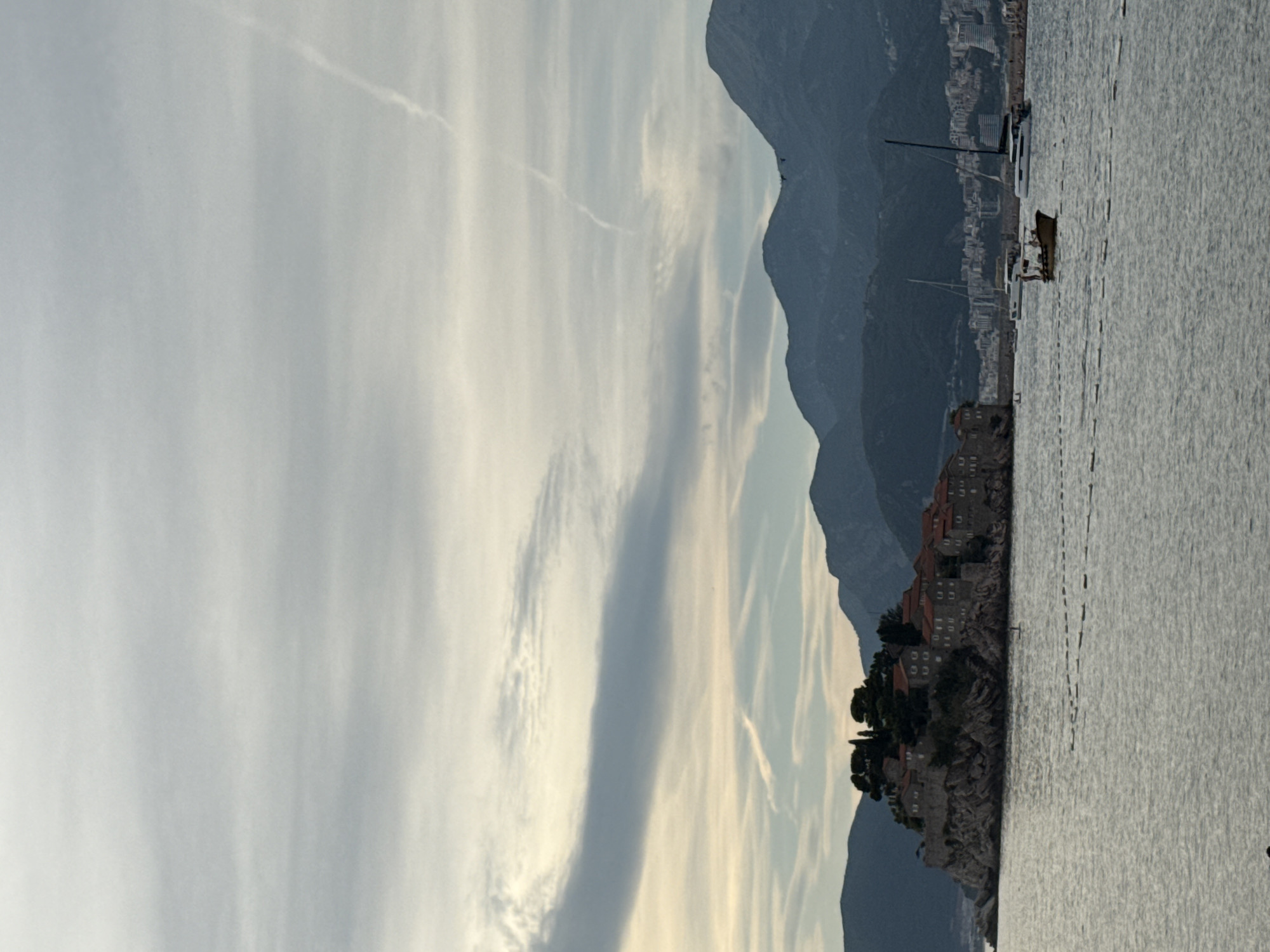 This image captures a serene coastal scene in Sveti Stefan, Montenegro. The foreground is dominated by the calm, rippling water of the Adriatic Sea, with a faint line of buoys marking a boundary in the middle distance. A small, yellow boat with a canopy and several figures aboard is navigating the water. To the left, a rocky island, fortified with stone buildings topped with red tile roofs and scattered trees, juts out of the sea. In the background, a range of lush, dark green mountains rises majestically against a sky filled with soft, layered clouds, some of which are streaked with faint white trails, possibly from aircraft. Further along the coast, a scattering of white buildings and yachts are visible, suggesting a developed area. The overall atmosphere is one of peaceful natural beauty and coastal charm.