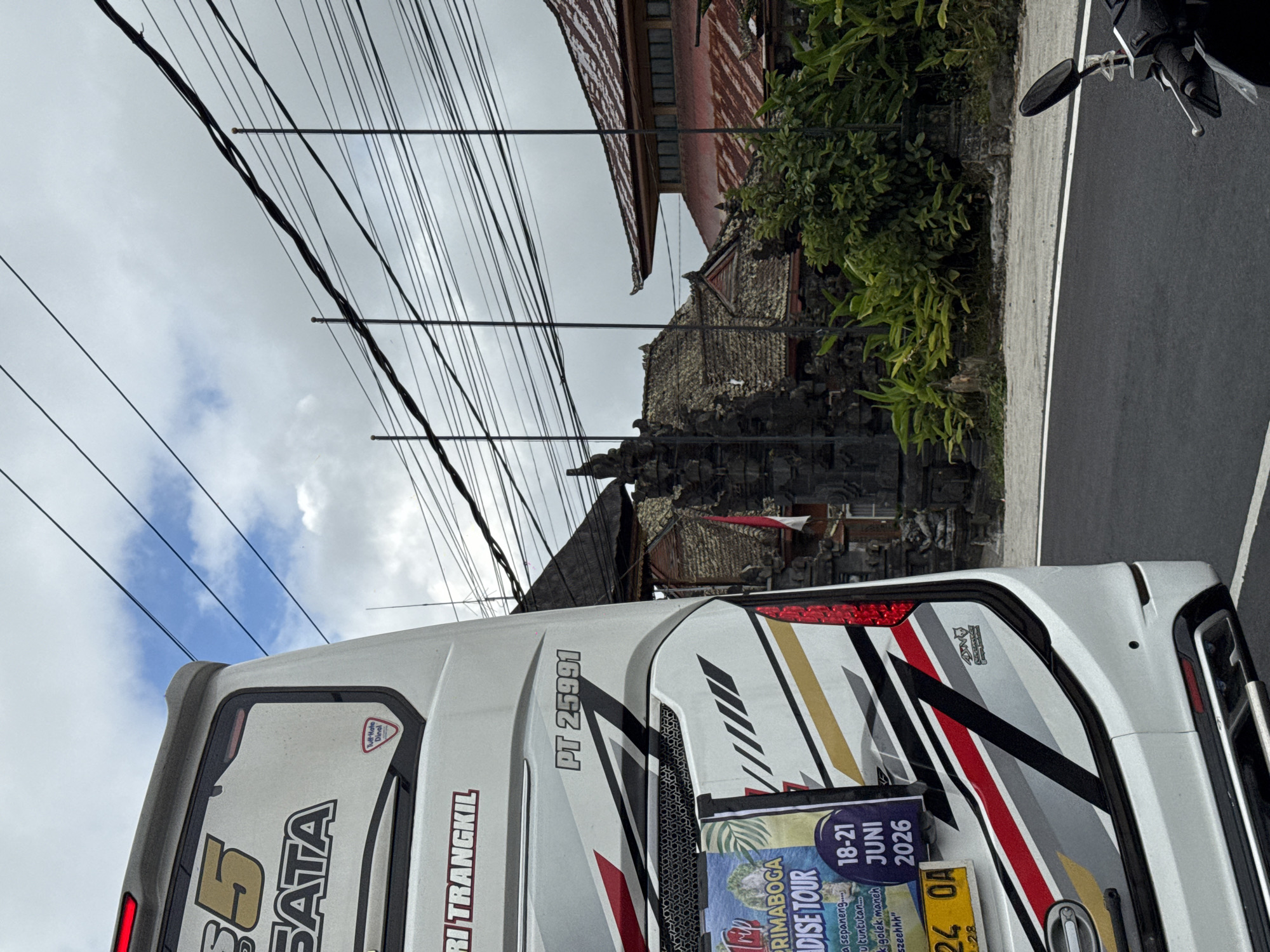In Kintamani, Indonesia, a white tour bus with red and gold accents is parked on a paved road. The bus has "PRIMABOGA PARADISE TOUR" written on a sticker. The license plate is visible and reads "24 QA". Behind the bus, a traditional stone gate with a red and white flag is visible, along with lush green foliage and buildings with terracotta roofs. Overhead, a network of power lines stretches across a cloudy sky. A motorcycle is parked on the right side of the road.