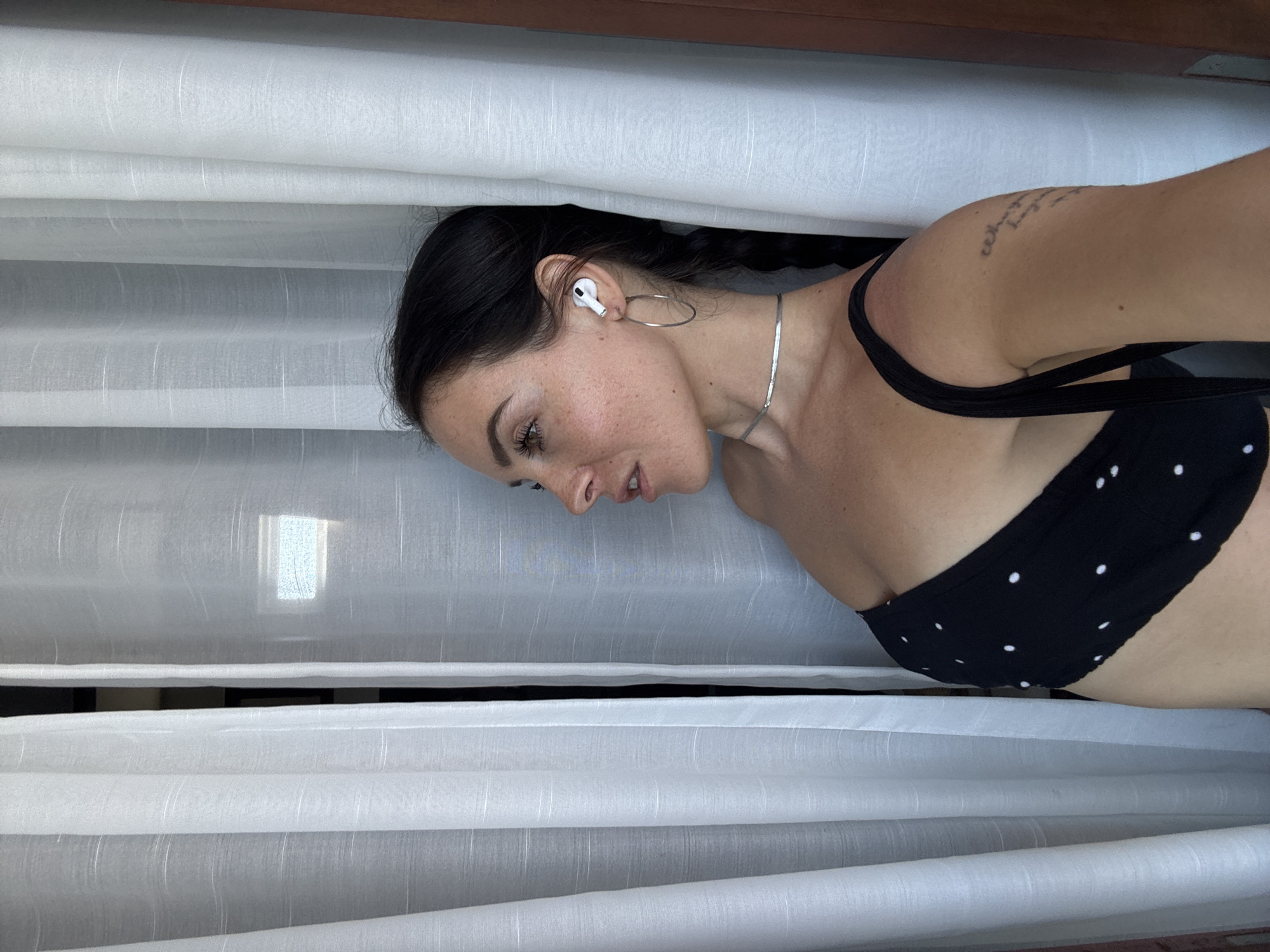 The media file shows a woman lying down, partially obscured by white curtains. She has dark hair styled in a braid and is wearing a black bikini with white polka dots. She has a silver necklace and is wearing white earbuds. A tattoo is visible on her upper arm. The scene appears to be indoors, with white curtains dominating the frame. Sunlight filters through the curtains, suggesting it is daytime. The woman's face is turned to the side, and her expression is neutral. The overall mood is relaxed and casual. The location context provided is North Kuta, Indonesia.