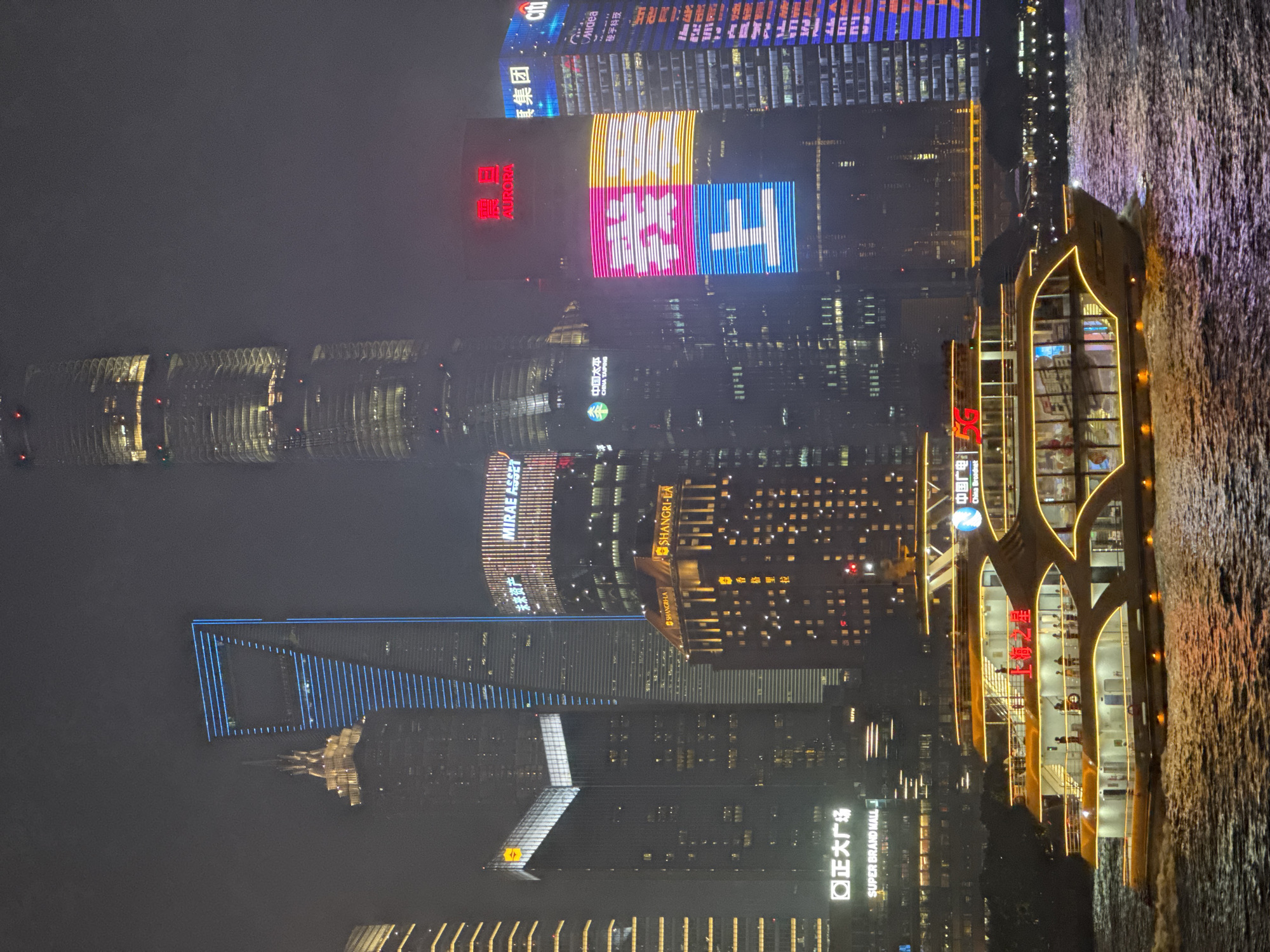 This is a nighttime aerial photograph of the Shanghai skyline, likely taken from a boat on the Huangpu River. The scene is dominated by towering skyscrapers adorned with vibrant neon lights and large illuminated signs. The water in the foreground reflects the city lights, creating a shimmering effect.
Prominent structures include the Oriental Pearl Tower, Shanghai Tower, and Jin Mao Tower, which are all illuminated in various colors. Signs visible on the buildings include "SUPER BRAND MALL," "MIRAE ASSET," "SHANGRI-LA," "CHINA TAIPING," "China Broadnet," "AURORA," "Midea," and "楼宇科技" (Building Technology). One building prominently displays a large, colorful light show with the characters "我上" (I'm up) and a stylized graphic. A large boat with illuminated windows and railings is visible in the foreground, suggesting a river cruise.
The sky appears to be hazy or foggy, softening the edges of the distant buildings and adding a moody atmosphere to the scene. The time of day is clearly night, with the artificial lights being the primary source of illumination. The overall impression is one of a bustling, modern metropolis at night, showcasing its impressive architecture and commercial signage.