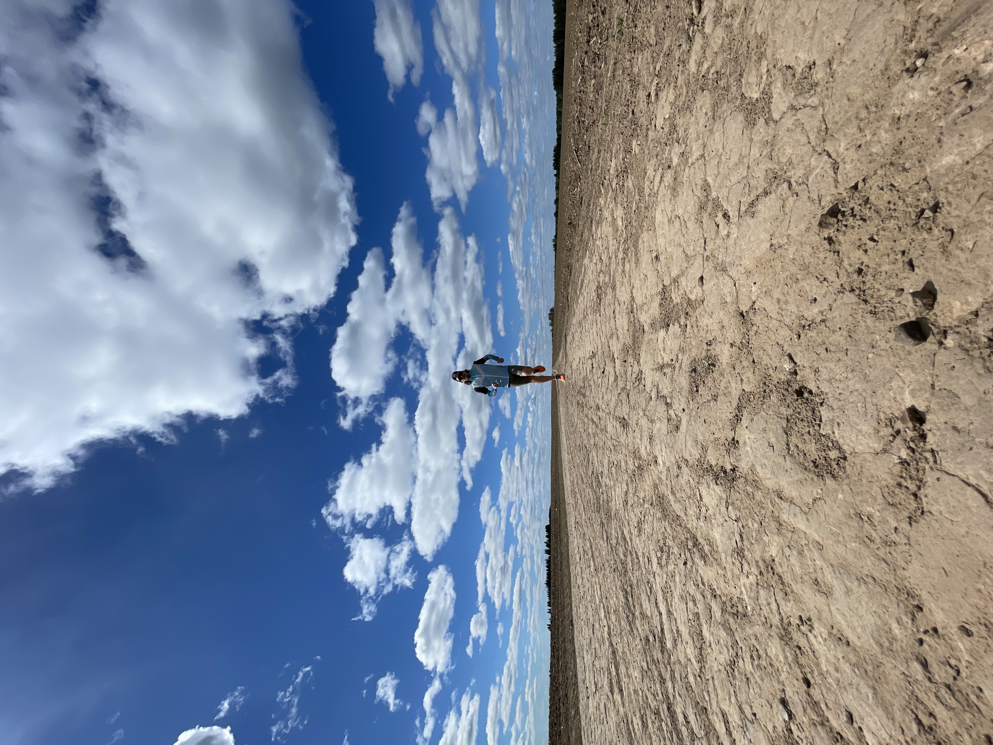 The image depicts an individual running in Perevoznoe, Russia. The scene is split horizontally, with a dry, cracked dirt field on the right and a vibrant blue sky with puffy white clouds on the left. A distant line of trees marks the horizon between the field and the sky. The runner appears to be wearing athletic clothing and is positioned in the center of the frame, suggesting a running or jogging motion. The perspective creates the illusion of a vast, open landscape.