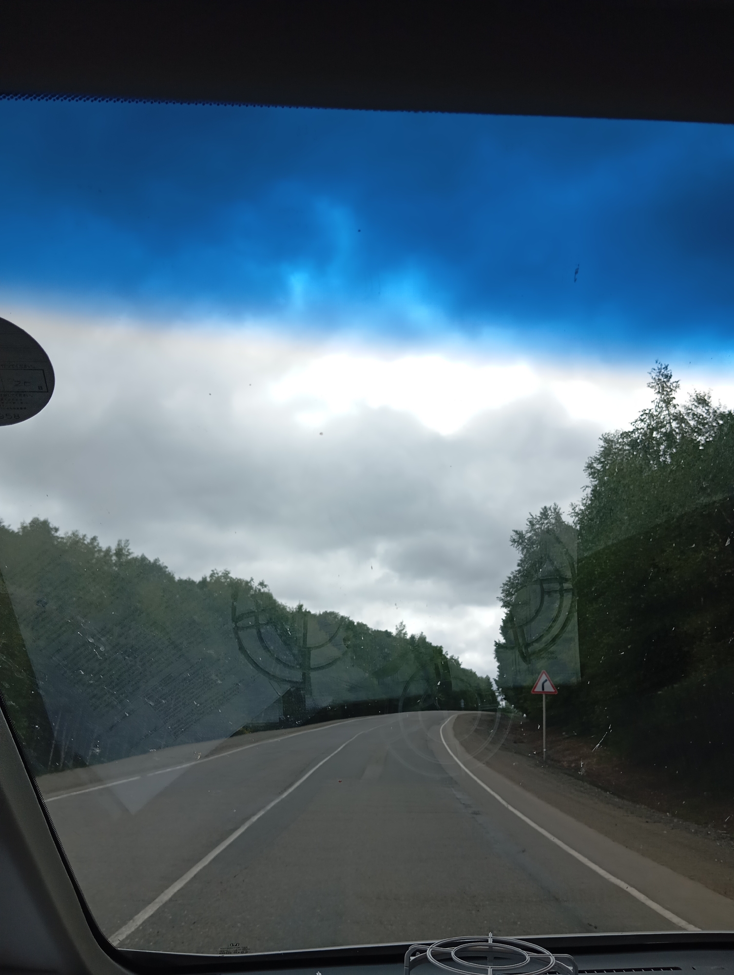 The image captures a view from inside a vehicle looking out through the windshield. The scene depicts a paved road winding through a forested area. The sky is overcast with patches of blue sky visible. On the right side of the road, a triangular traffic sign with a black arrow indicating a curve to the right is present. The windshield has visible smudges and streaks, indicating it is likely dirty. The surrounding environment is characterized by dense green trees lining the road. There are no people or animals clearly visible in the image. The overall impression is that of a journey through a rural, wooded landscape.
