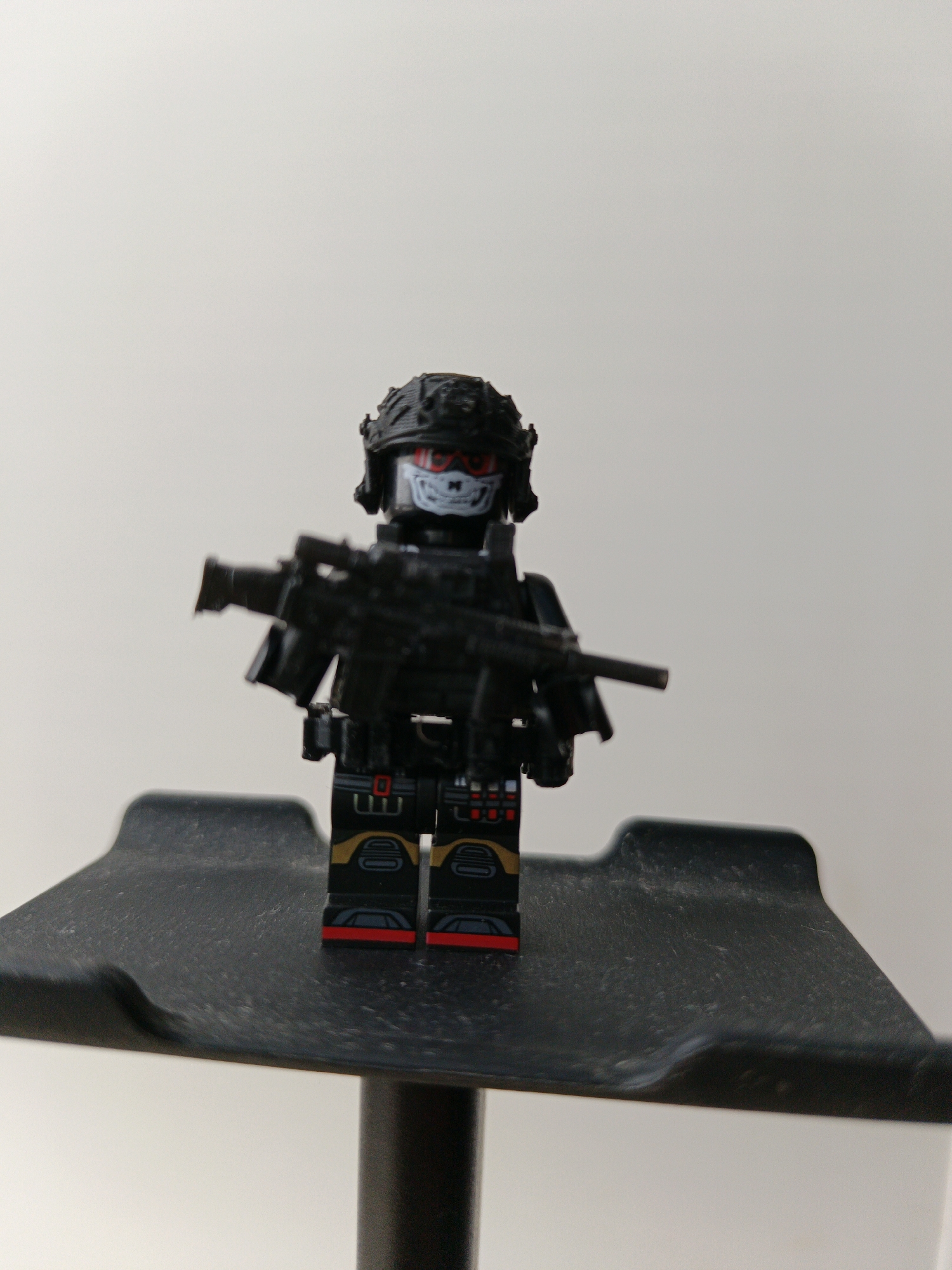 A Lego minifigure resembling a tactical soldier stands on a black surface. The figure has a helmet, a skull-printed face, and is holding a black rifle. The background is a plain, light color.