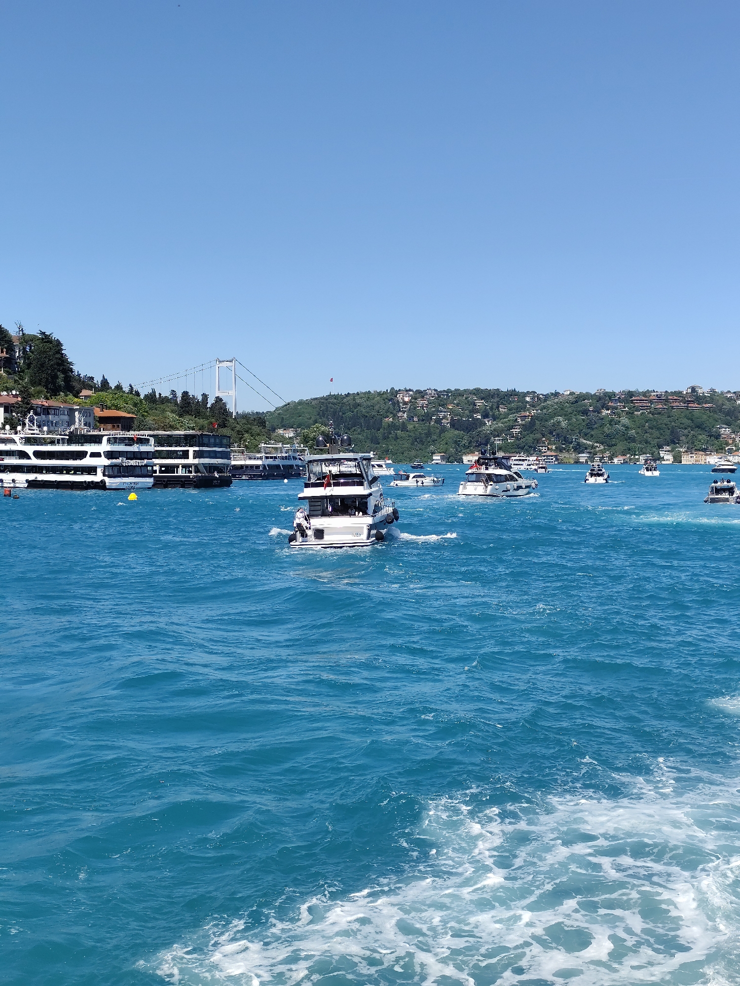 The image captures a bustling maritime scene in Beşiktaş, Turkey, characterized by numerous boats navigating through turquoise waters. In the foreground, churning water reveals the wake of the photographer's vessel. Further out, a variety of boats, including ferries and yachts, traverse the waterway. One prominent yacht is centrally positioned in the midground. In the background, the iconic Bosphorus Bridge is visible, connecting the European and Asian sides of Istanbul. The landscape includes verdant hills dotted with residential buildings, typical of the urban landscape in Beşiktaş. The sky is clear and blue, indicating a sunny day. A yellow marker buoy floats in the water near the ferries on the left. A Turkish flag can be seen on top of the hill on the right of the image.
