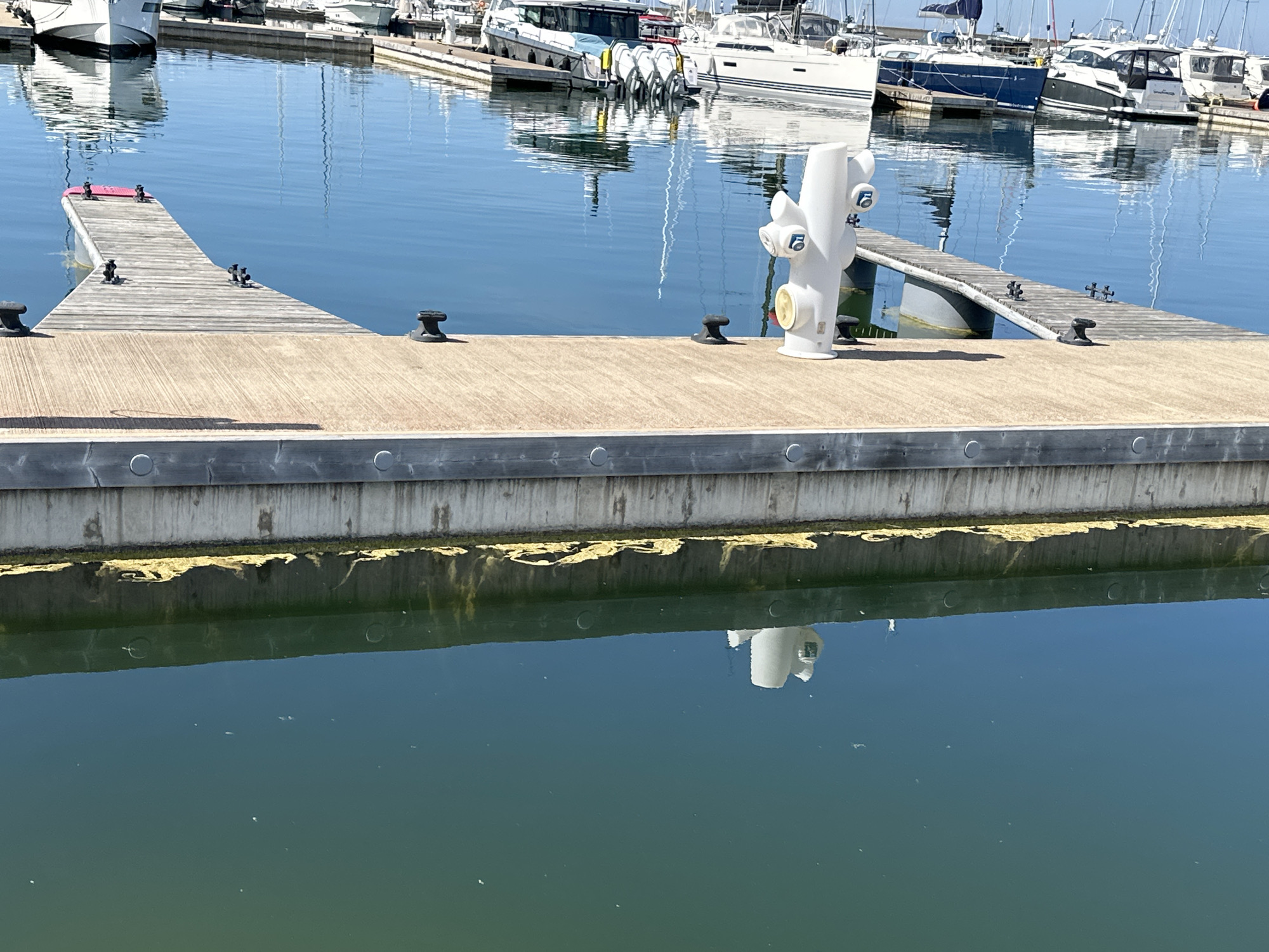 This image depicts a marina in Tallinn, Estonia, on a clear, sunny day. In the foreground, a light-colored wooden dock stretches horizontally across the frame, equipped with several black mooring cleats. A white, multi-outlet utility pedestal, featuring numerous blue-capped and two yellow-capped ports for water or electricity, stands on this main dock. To the left, a narrower, darker wooden finger pier extends diagonally into the water, also fitted with cleats and ending in a red-capped section.
The calm, blue water reflects the distant boats and structures. In the background, an assortment of recreational boats and yachts, predominantly white with some blue accents, are moored along other piers, their hulls and masts clearly mirrored in the water's surface. Below the foreground dock, the water appears a darker greenish-blue, and a distinct band of yellowish-green marine growth is visible along the concrete retaining wall just above the waterline. The white utility pedestal's reflection is clearly visible in this darker water. No individuals are present in the scene.