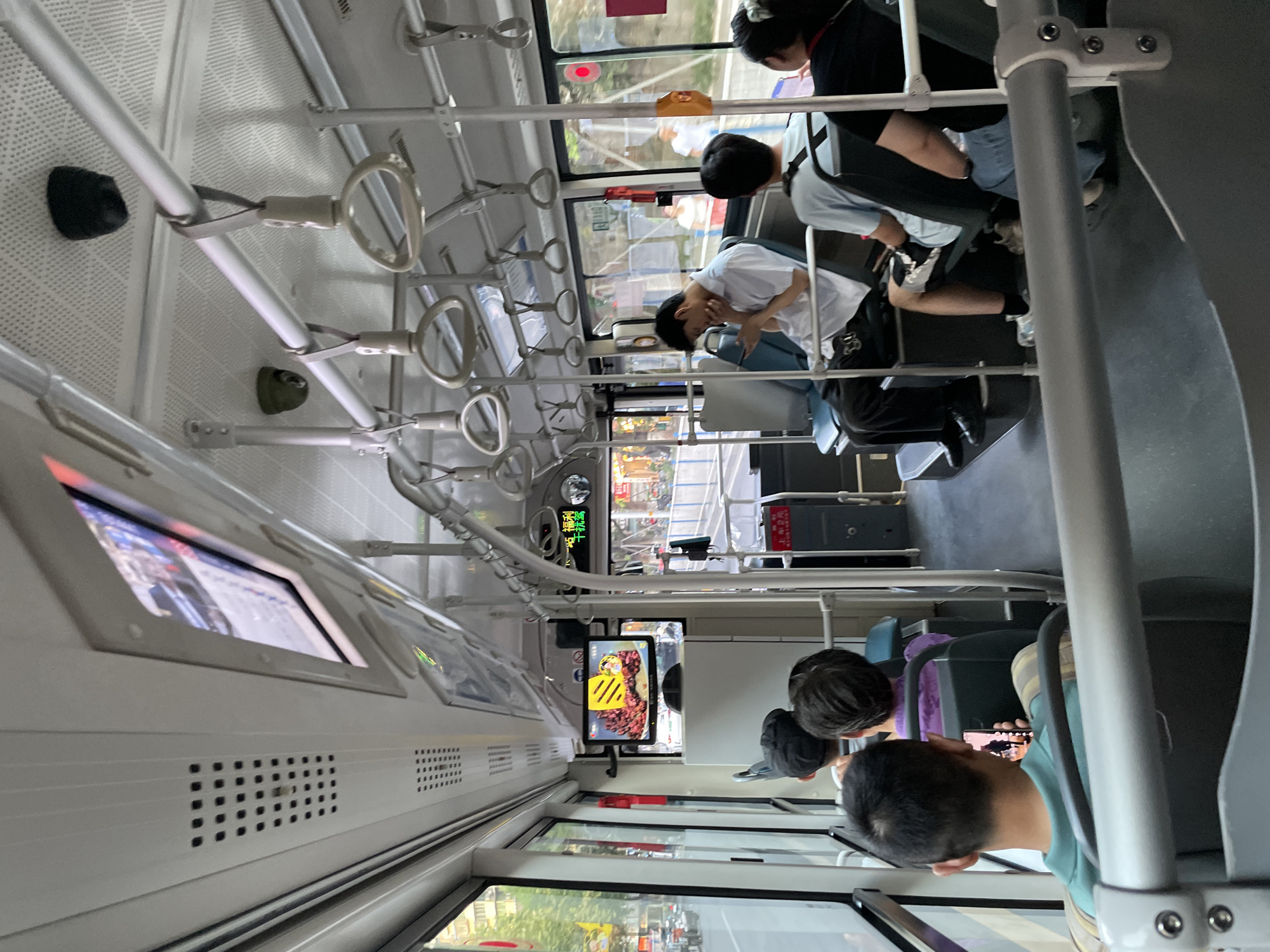 The image depicts the interior of a bus, possibly in Nan'an District, China. Several passengers are seated on the bus, some looking at their phones or the screens mounted overhead. One passenger in the foreground is looking at their phone, while another in the middle is also engaged with a device. The bus has various handrails and overhead compartments. A screen at the front of the bus displays an image of food. Outside the windows, a street scene with buildings and traffic is visible. The lighting suggests it is daytime.