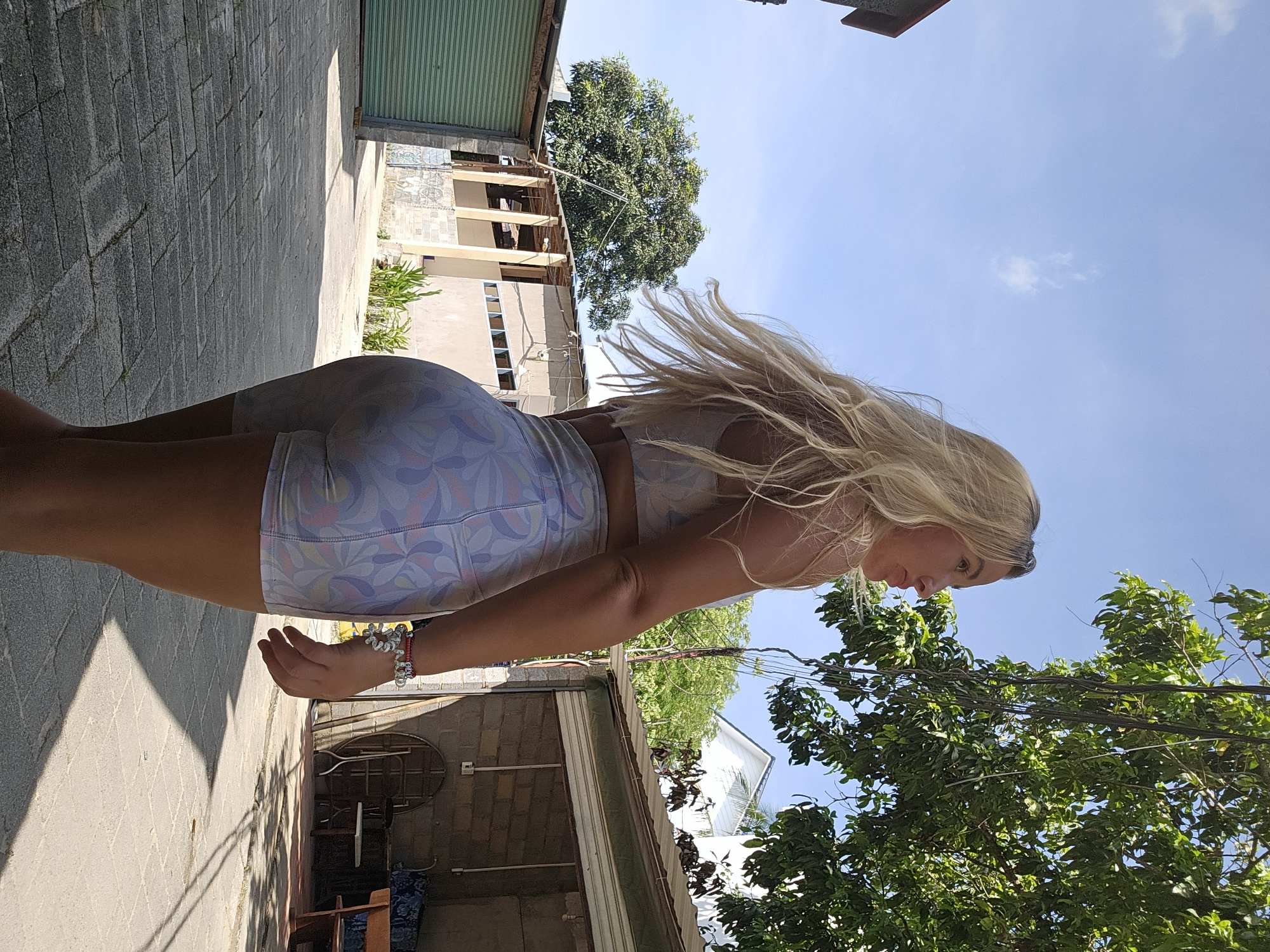 A blonde woman is standing on a paved walkway outdoors in what appears to be a residential or industrial area. She is wearing a light-colored patterned sports bra and matching shorts. Her hair is long and wavy, and she is looking to her right. Behind her, there are buildings, trees, and a blue sky with a few clouds. The lighting suggests it is daytime and sunny, with shadows visible on the ground. The location is identified as Phu Thuy Ward, Vietnam.