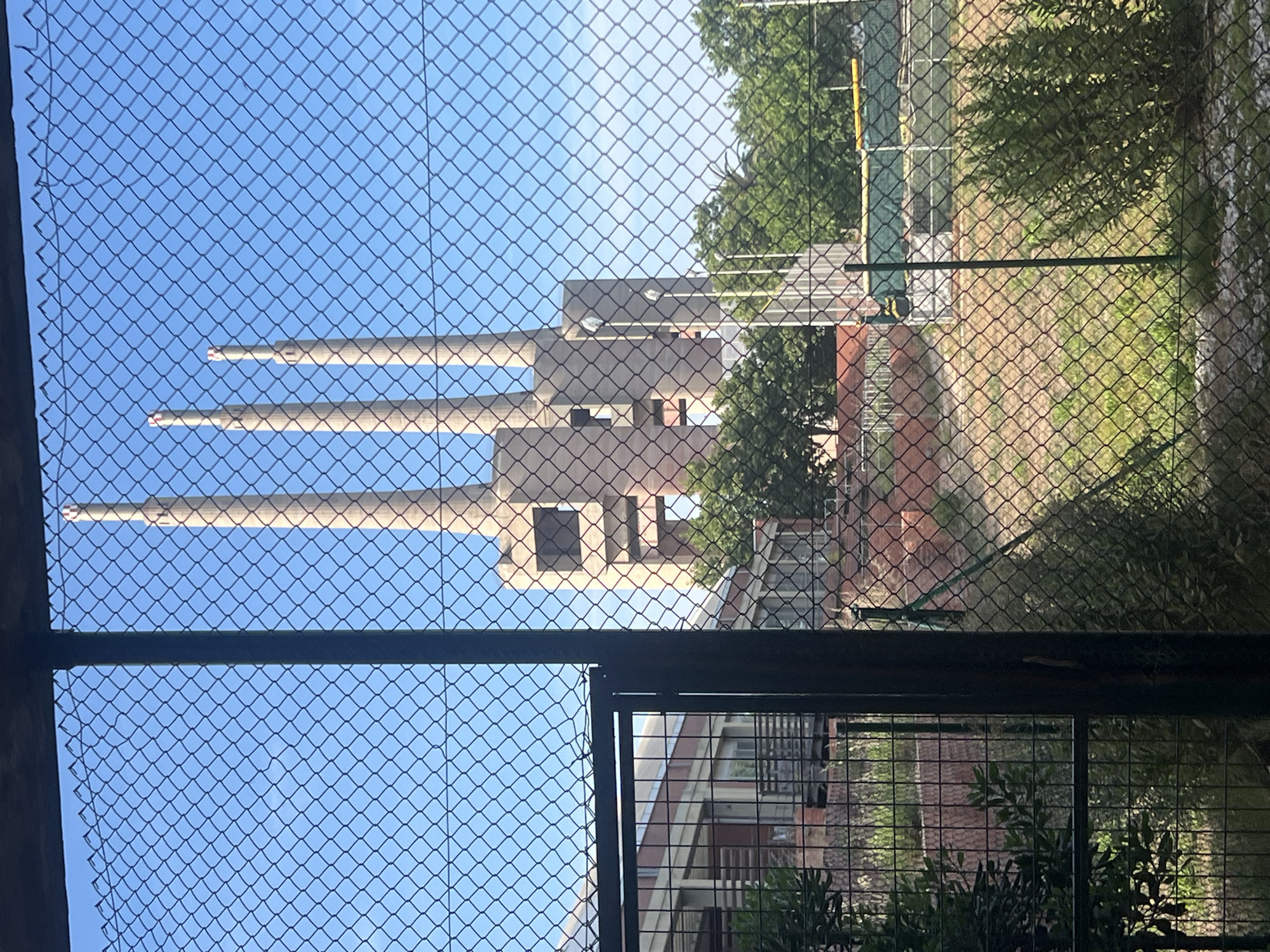 The image captures a view through a chain-link fence of the three chimneys of the Sant Adrià de Besòs thermoelectric power station, located in the city of Sant Adrià de Besòs, Spain. The chimneys are the dominant feature in the background, silhouetted against a clear blue sky. They are cylindrical and made of concrete, with a reddish-brown band near the top of each. In the middle ground, there are trees and the upper structures of the power station. The foreground is dominated by the chain-link fence, with a dark metal gate visible on the left. Beyond the fence, there is a grassy area with some sparse vegetation, and further in the distance, it appears to be a sports field with a yellow barrier. There are no people visible in the image. The overall scene suggests an industrial area bordering a recreational space.