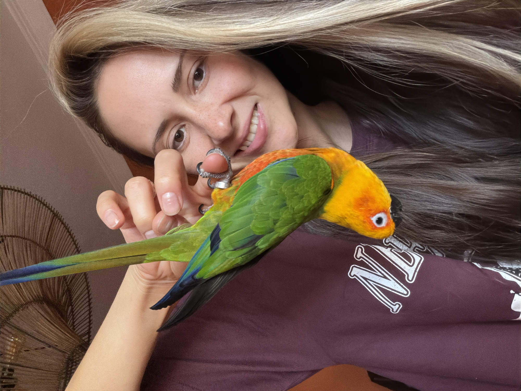 A woman with long, partly blonde hair, freckles, and a smiling expression is holding a colorful parrot. The parrot has a bright orange head, green back and wings, and a blue tail. The woman is wearing a dark red t-shirt with white lettering. A decorative fan can be seen in the background. The location is not discernible as Khimki, Russia based on the image.