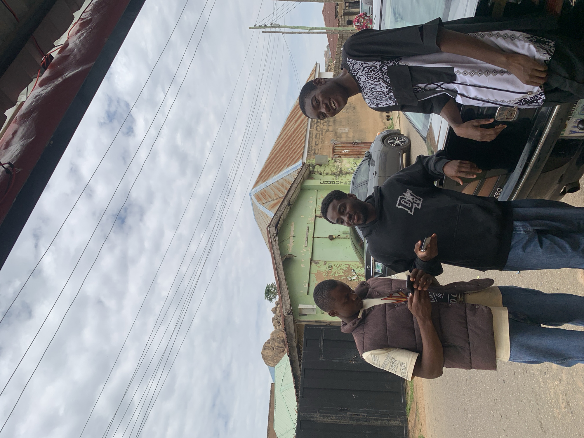 Three young adult males are depicted standing on a paved street in Jos, Nigeria, under an overcast sky filled with dense, light grey clouds. Numerous black utility wires traverse the upper portion of the frame.
The man on the left, with dark skin and short dark hair, is wearing a light-colored t-shirt, a dark brown puffer vest, and blue jeans. He is looking down, holding and interacting with a black smartphone with both hands.
The middle man, also with dark skin and short dark hair, sports a light beard. He is dressed in a black hooded sweatshirt featuring a white "D" emblem and blue jeans. He holds a silver smartphone in his left hand and is observed leaning his right arm on the trunk of a black sedan, looking towards the right with a slight smile.
The man on the right, dark-skinned with short dark hair, is wearing a black and white patterned tunic-style shirt and dark trousers. He is smiling broadly, looking directly at the viewer, with his left arm resting on the trunk of the same black sedan.
The black sedan, positioned behind the middle and right men, has a partially visible Nigerian license plate at the bottom which reads "ABUJA". A grey sedan is parked further down the street behind the middle man.
In the mid-ground, a single-story building with faded light green paint and a rusty corrugated iron roof is prominent. Vertical text "COMPUTER" is visible on its left side. Behind this structure, significant rock formations, characteristic of the Jos Plateau, are visible. Another building with a steeply pitched, very rusty corrugated iron roof is also present. A black metal gate and fence are visible on the far left. A wooden utility pole stands on the right side of the frame. A red, textured material, possibly an awning or part of a building structure, is visible at the very top of the image. The ground is a paved surface, indicating a street or pathway.