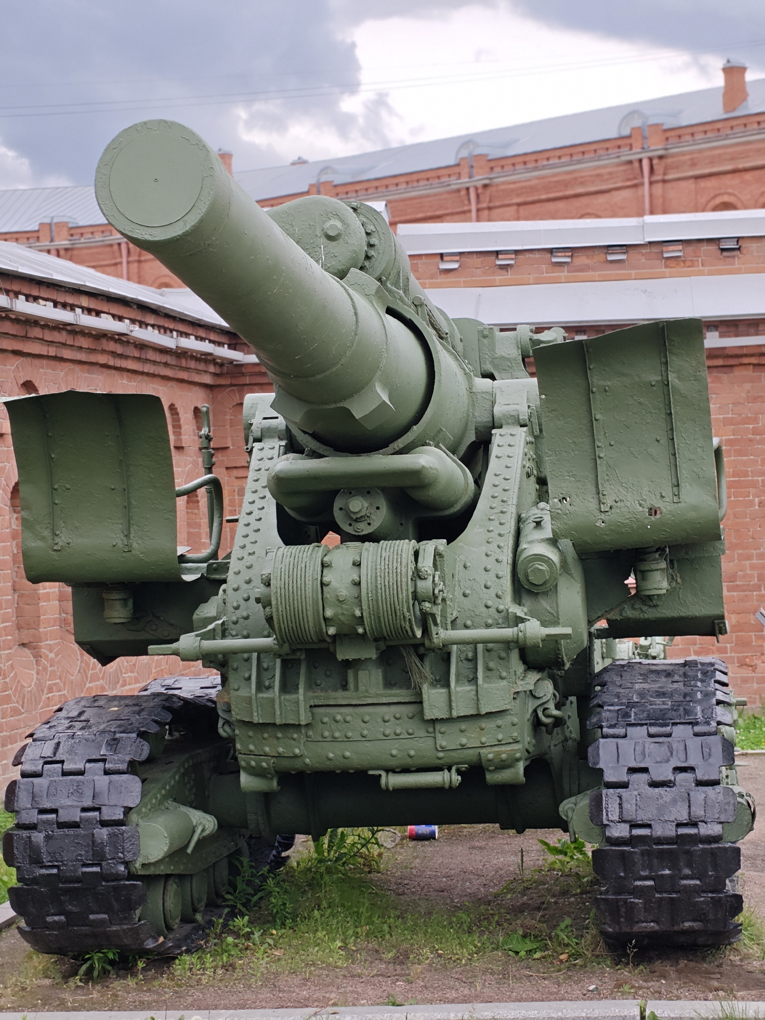 An outdoor, medium-shot, eye-level photograph shows a large, olive-green, tracked artillery piece positioned in front of a red brick building with a light gray roof and a cloudy sky overhead. The artillery piece, likely a self-propelled gun or howitzer, is a complex mechanism with a prominent barrel extending to the left of the frame. The barrel is attached to a wheeled carriage with a visible winch mechanism on the front. Large, black rubber tracks are on either side of the carriage, resting on a patch of grass and gravel. The red brick building in the background appears to be a historic structure, possibly part of a museum or fortification. The overall impression is of a static military display in an urban environment. The location is the Peter and Paul Fortress in Saint Petersburg, Russia.