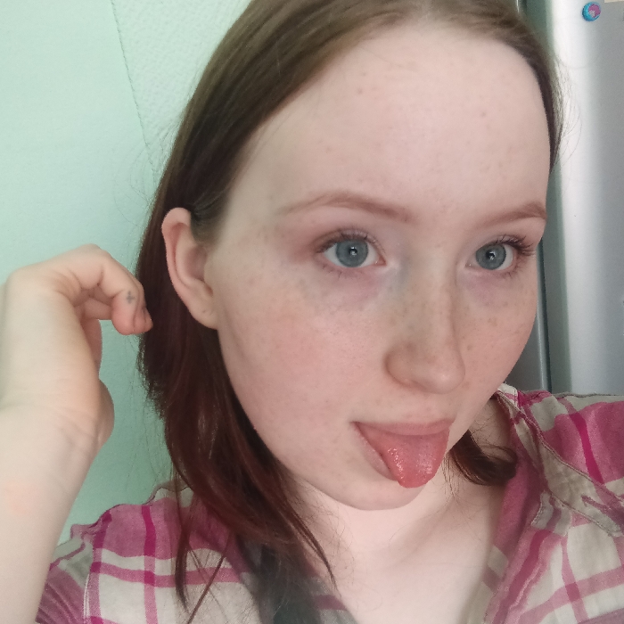 The image displays a head-and-shoulders portrait of a young adult female in an indoor, domestic setting. The precise location within Chaikovsky, Russia, is not discernable from the visual information. The subject has fair skin adorned with numerous freckles, light reddish-brown hair, and blue eyes. She is wearing a plaid shirt featuring white and pink patterns. Her left hand is raised to the side of her head, with a small, dark, cross-shaped tattoo visible on the lateral surface of her left pinky finger. Her mouth is open, with her tongue extended outwards and slightly to the left. The background on the left consists of a smooth, light mint green wall, while the background on the right exhibits a reflective, metallic surface, likely a household appliance.