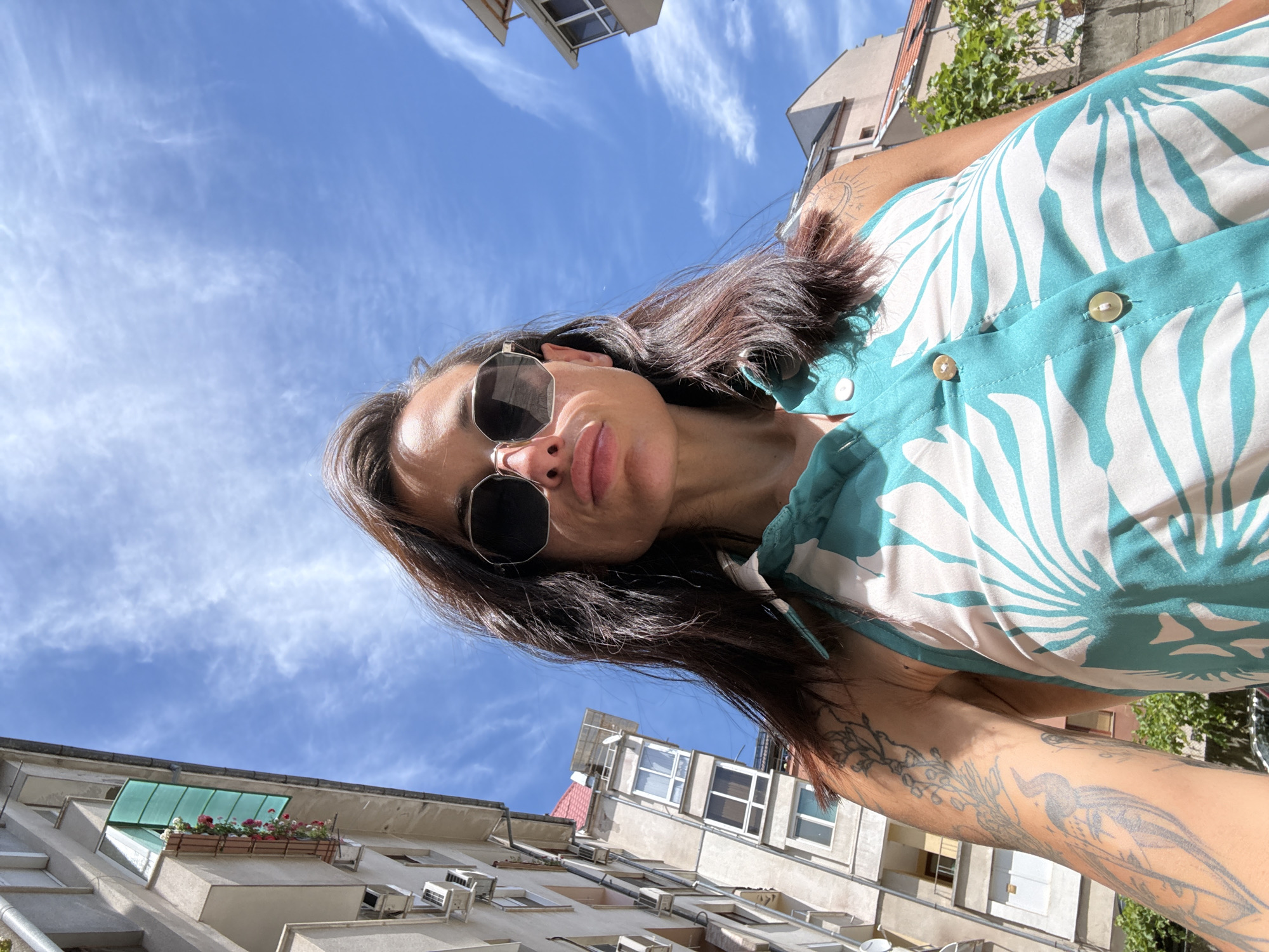 A woman is taking a selfie outdoors, looking up at the camera with a slight smile. She is wearing sunglasses and a sleeveless teal and white floral-patterned top. Visible tattoos adorn her arm. The background consists of a bright blue sky with wispy white clouds. Below the sky, the tops of buildings are visible, with some showing balconies with flower boxes and air conditioning units. The scene appears to be set in an urban environment, possibly in Pleven, Bulgaria, on a sunny day. The upward angle of the shot suggests the woman is holding the camera above her head. The lighting indicates it is daytime, likely late morning or afternoon, given the strong sunlight.