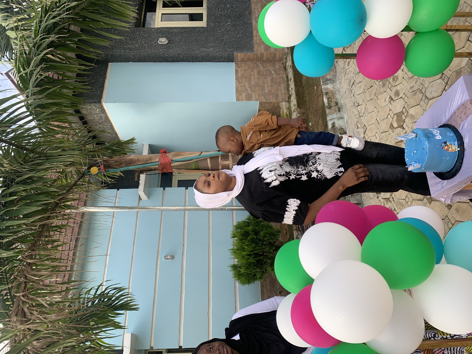 The image depicts an outdoor scene in the city of Jos, Nigeria, featuring three individuals and celebration decor in front of a building.
On the left, an adult female is partially visible, wearing a black head covering and a white top, looking towards the camera with a neutral expression. In the center, an adult female, wearing a white headscarf and a black t-shirt with white graphics, holds a young male child on her left hip. She is facing slightly to her right, with her gaze directed towards the viewer's left. The child, a toddler, wears a brown long-sleeved shirt with "AFRICHTU" (or similar text) on the shoulder, blue jeans, and white sneakers with "SPORTS" branding. The child's face is turned away from the camera.
In the foreground, large clusters of inflated balloons are present, primarily in white, green, pink (magenta), and light blue (cyan). A small, light-blue cake decorated with white elements and "Bluey" characters is situated on a black stand atop a small table draped with light purple fabric, positioned to the right of the central woman.
The background consists of a two-story building. Its upper section is painted light blue with three prominent white horizontal bands, and a cylindrical outdoor light fixture is mounted on the wall. The right portion of the building displays a darker, textured grey finish, with brown rectangular tiles along its lower section. Light-framed windows are visible. Large palm fronds extend into the frame from the top and left, partially obscuring the building and sky. A slender metal pole stands vertically in the center-right, and a wooden pole with red fabric tied around its base is on the far left. A small, green ornamental bush is located in the mid-ground between the two women. The ground is paved with light-colored, interlocking patterned tiles, suggesting a patio or walkway. The scene is illuminated by natural daylight.