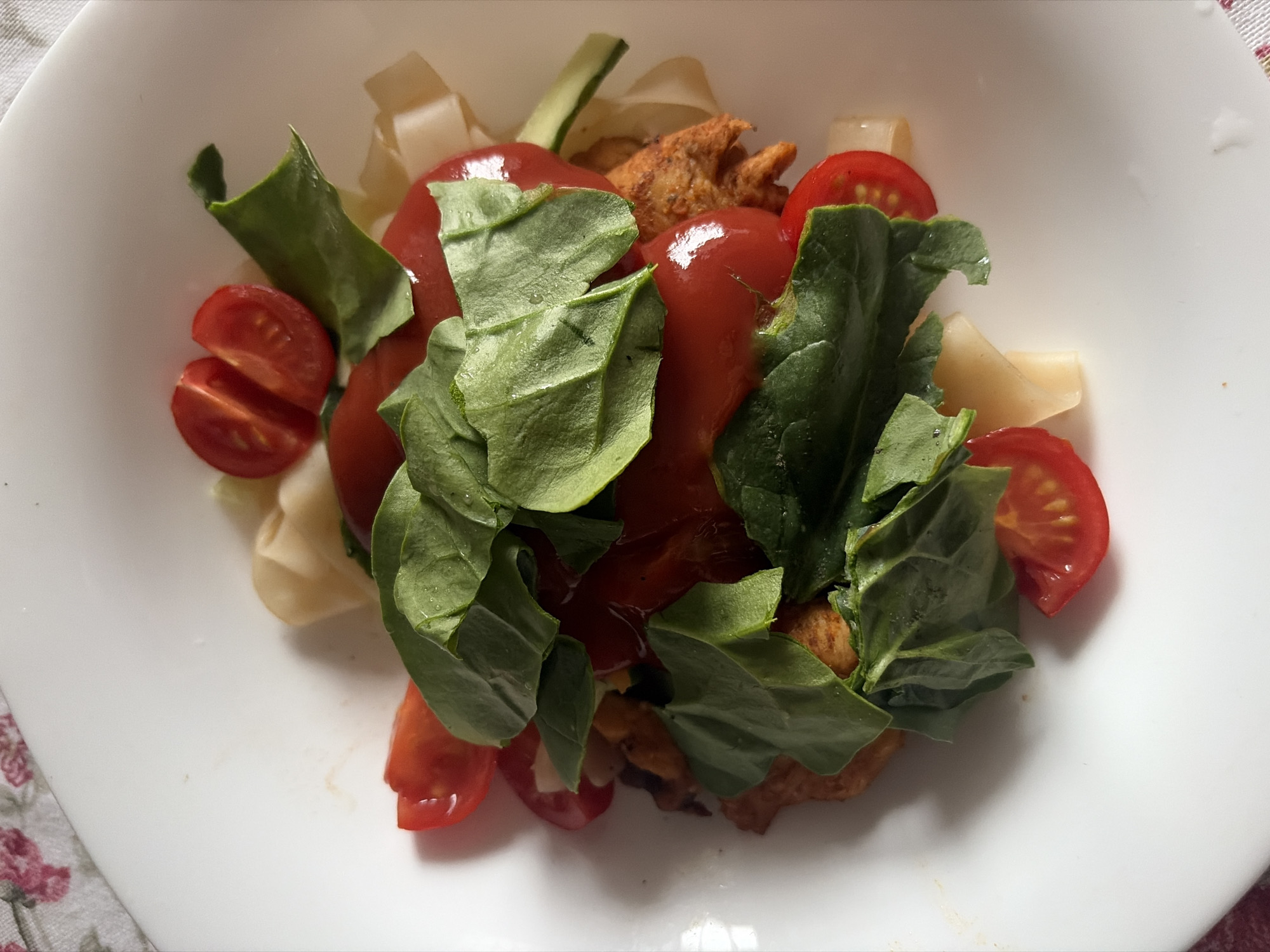 A top-down shot shows a white plate containing a meal of what appears to be a salad-like composition. Visible components of the meal include halved cherry tomatoes, leafy greens (likely spinach or a similar variety), pieces of cooked chicken, light-colored strips resembling noodles or perhaps cut vegetables, and a red sauce that appears to be tomato-based. The food is arranged in a central pile on the plate. Part of a tablecloth or surface with a floral pattern is visible in the lower left corner of the frame. The scene gives no indication of the location in Saint Petersburg, Russia.