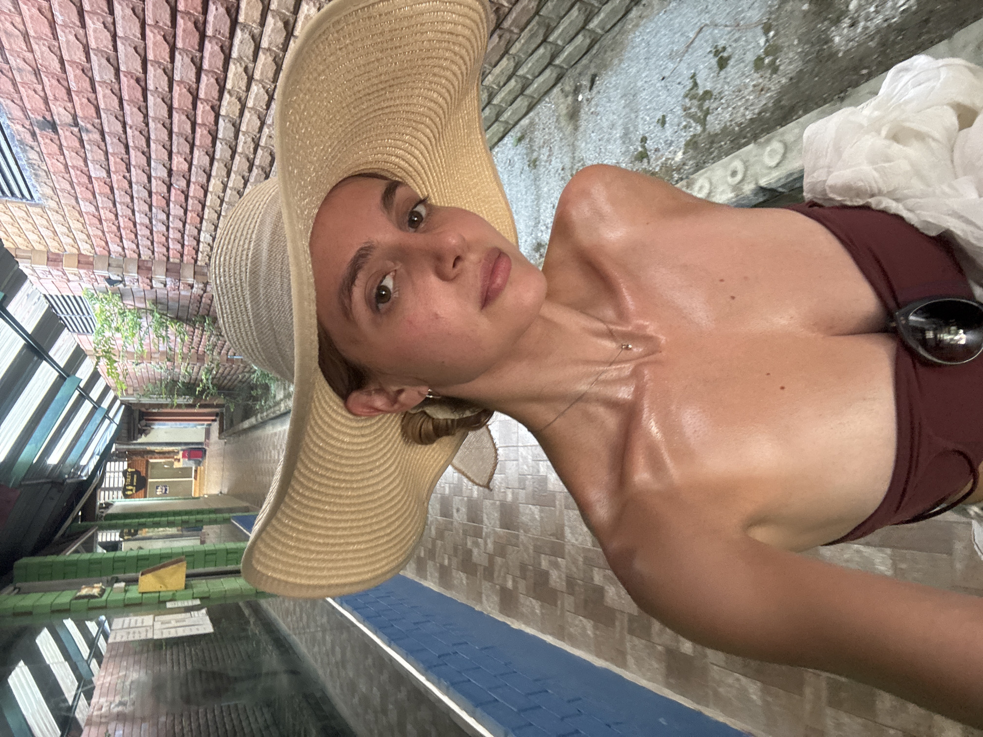 A woman with fair skin, wearing a large straw hat and a maroon bathing suit, is posing for a picture. The background features what appears to be a therapeutic pool setting. There are multiple pools with green edges, and what looks like reflective windows are visible, suggesting an indoor location. The setting is consistent with the Ban Khlong Khian area of Thailand, known for its wellness spas. The woman's skin is shiny, potentially from water or lotion. She is also wearing a necklace. Sunglasses are tucked into the top of her bathing suit. A white cloth is draped over one shoulder. The perspective of the photo suggests she is holding the camera at an angle. The architectural style of the surrounding walls is brick-like and lined.