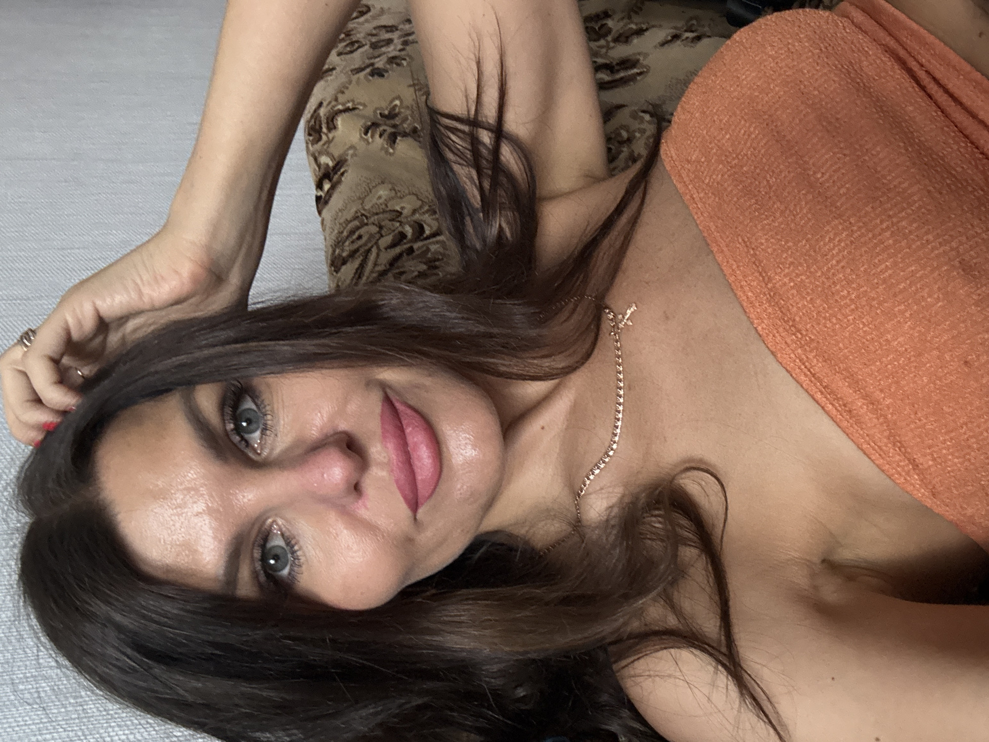 A close-up selfie shows a woman with dark brown hair, blue eyes, and light skin. She is wearing an orange strapless top and a gold necklace with a cross pendant. Her right arm is raised, with her hand resting on her head. She is looking at the camera with a slight smile. The background appears to be a patterned couch or chair. There is no information about the location in Dubovaya, Russia.