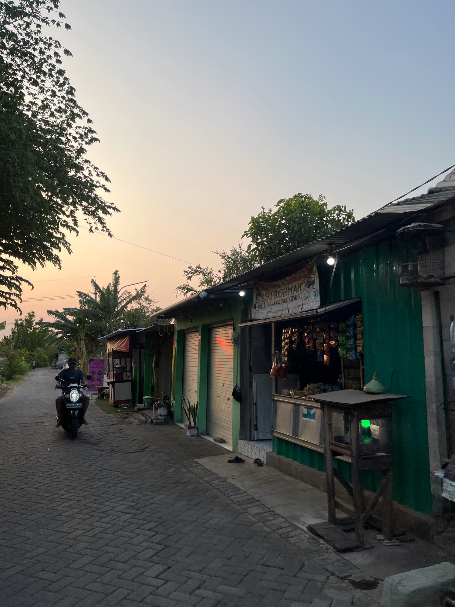 In Sruni, Indonesia, at dusk, a person rides a scooter down a paved road lined with small storefronts. The sky is a soft gradient of orange and pale blue, indicating the setting sun. The storefronts, constructed with corrugated metal walls painted green, have open fronts displaying various products on shelves. One storefront has a banner that reads "IBU ROHMAH (CABANG MAK KIRAH)". A birdcage is visible on a post outside one of the shops. The road is paved with interlocking dark grey bricks. Trees and lush green foliage border the road. A few potted plants are placed near the storefronts.