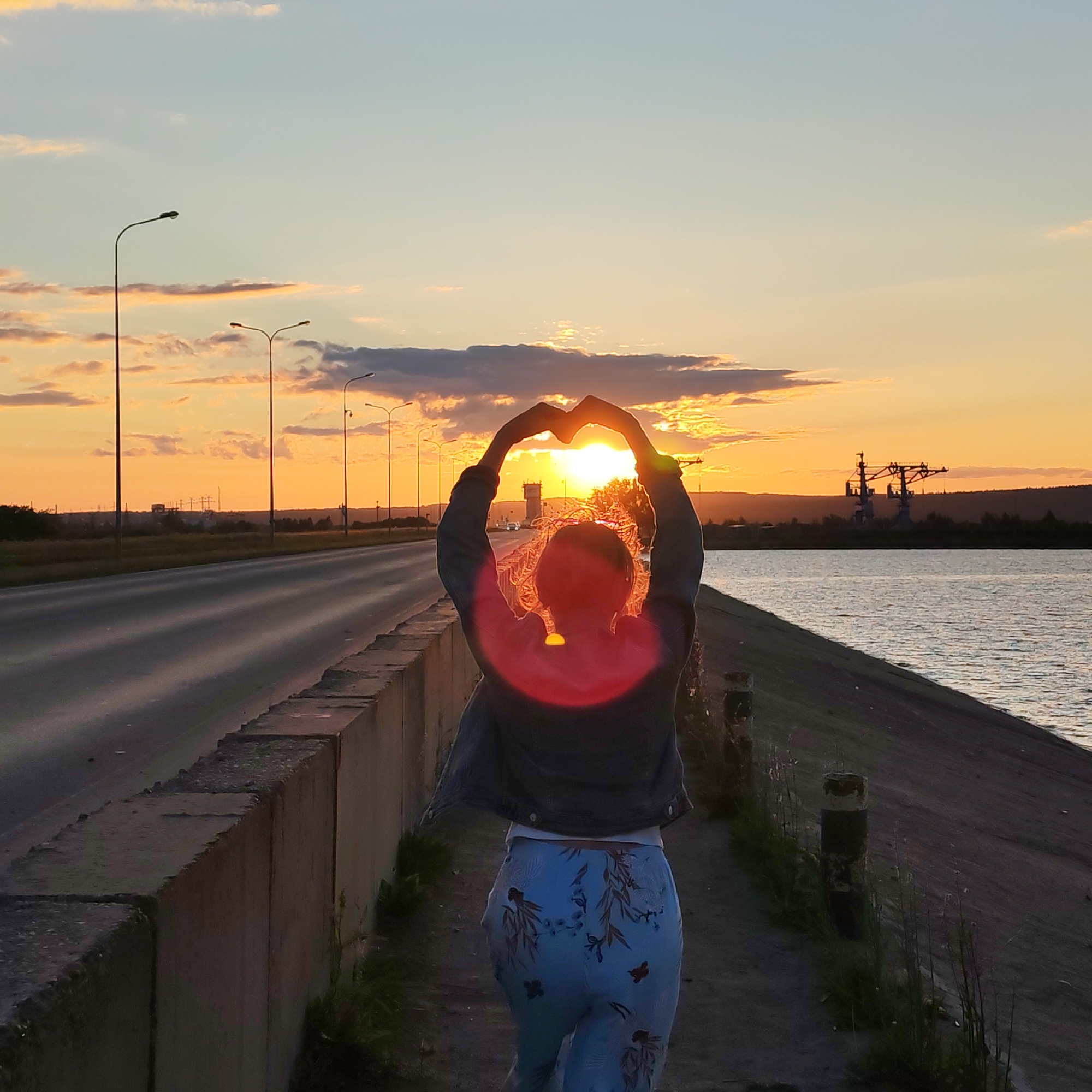 The image shows a person silhouetted against a sunset on a dam overlooking a body of water. The person is standing on the dam, facing away from the camera, with their arms raised and hands forming a heart shape around the setting sun. The sun is a bright orange disc with rays extending outwards, partially obscured by clouds. The sky is a gradient of blue and orange.
To the left of the person is a paved road with streetlights. In the distance, industrial cranes are visible against the horizon. The dam itself is made of concrete, with some grass growing along its edge. The water on the right appears calm, reflecting the colors of the sky. The overall atmosphere is serene and picturesque.
This scene is likely located on the dam of the Votkinsk Hydroelectric Power Station in Chaikovsky, Russia, which is a prominent feature of the city and a popular spot for scenic views.