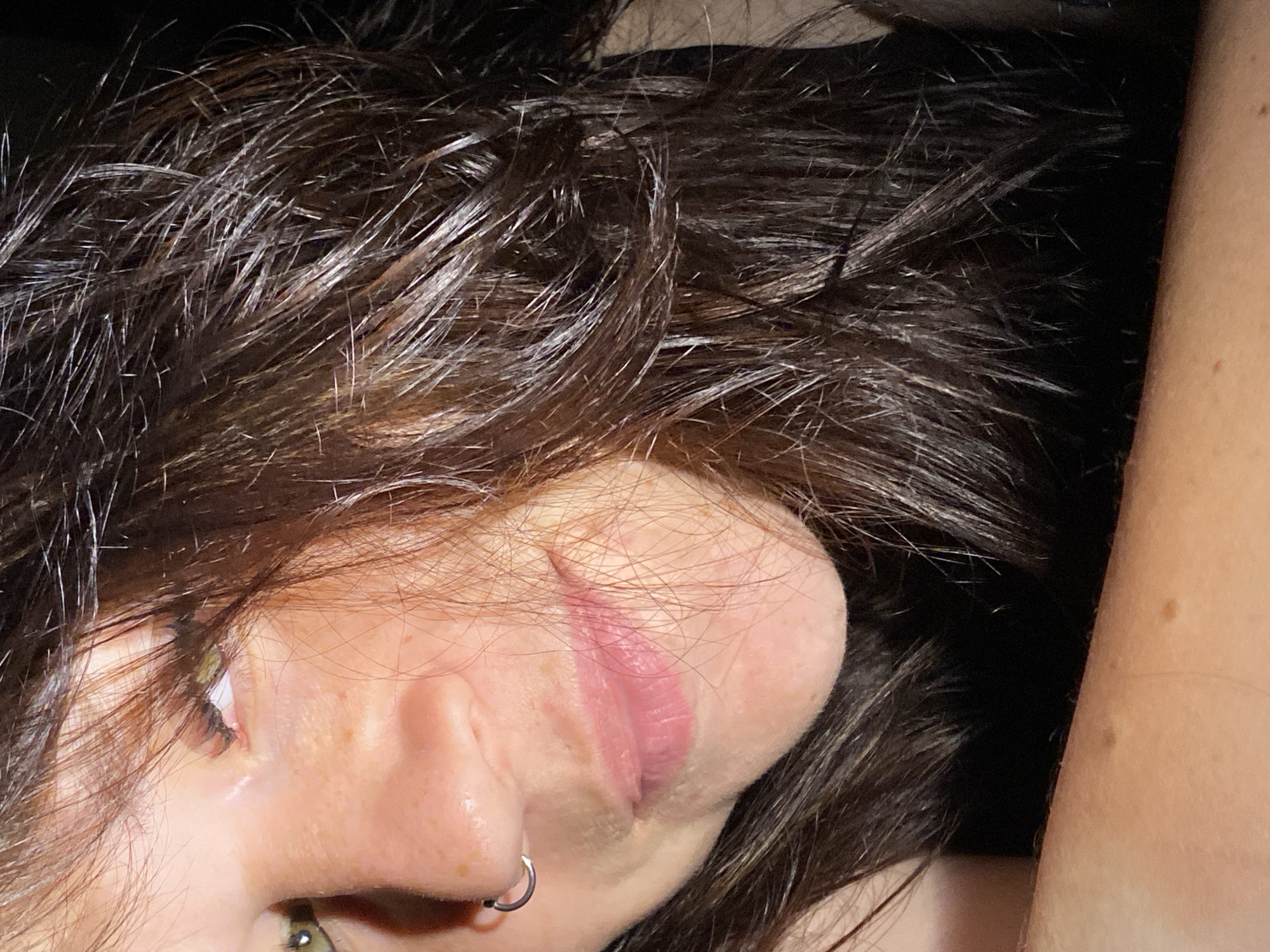 The image is a close-up, self-portrait of a person with dark, slightly messy hair and fair skin. The person has green eyes and a nose ring. Part of their face, including their eye, nose, and mouth, is visible. Their lips are slightly parted. The background is dark. There is no discernible city or location information within the image itself or any surrounding context provided.