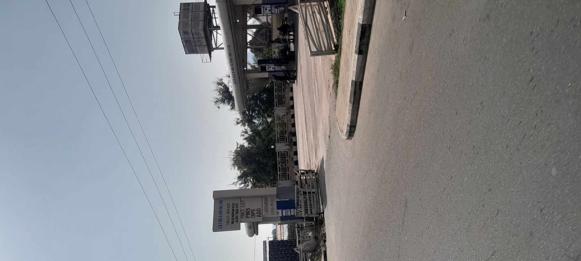 The image shows a gas station in Jalingo, Nigeria. The gas station is branded "A.K.S OIL & GAS NIG. LTD." A signpost lists "HEAD OFFICE MILEGO NUMAN ROAD JALINGO TARABA STATE", a "PRICE LIST" for PMS, DPK, and AGO, and mentions "LUBRICATION MINI-MART." The gas station features several fuel pumps under a canopy. A water tank is visible on a metal structure behind the canopy. Several people are present, some standing near the pumps and others sitting at a table. The scene is captured from a street-level perspective, with a paved road in the foreground. The sky is clear and blue.
