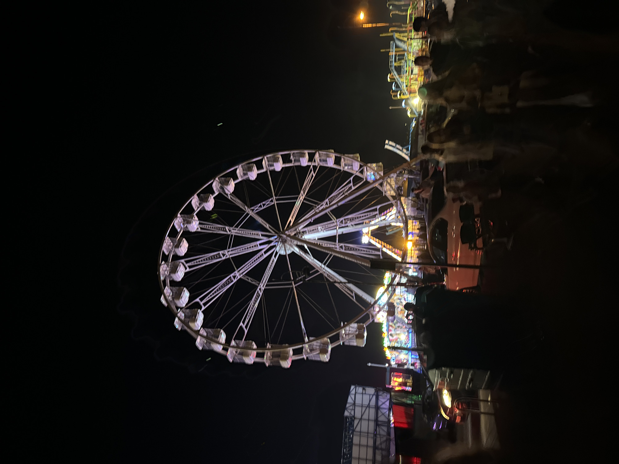 An outdoor nighttime scene at a fairground in Tramore, Ireland, features a large Ferris wheel with white gondolas illuminated by purple lights, dominating the center of the frame. To the left of the Ferris wheel, a tall, white, scaffolding-like structure with red lights is visible. The background is filled with other fairground attractions, including roller coasters and smaller rides, all illuminated with colorful lights against the dark sky. In the foreground, several cars are parked, and a group of people, some appearing blurred due to motion, are visible standing and walking. The overall atmosphere suggests a lively event with people enjoying the fair rides and attractions.