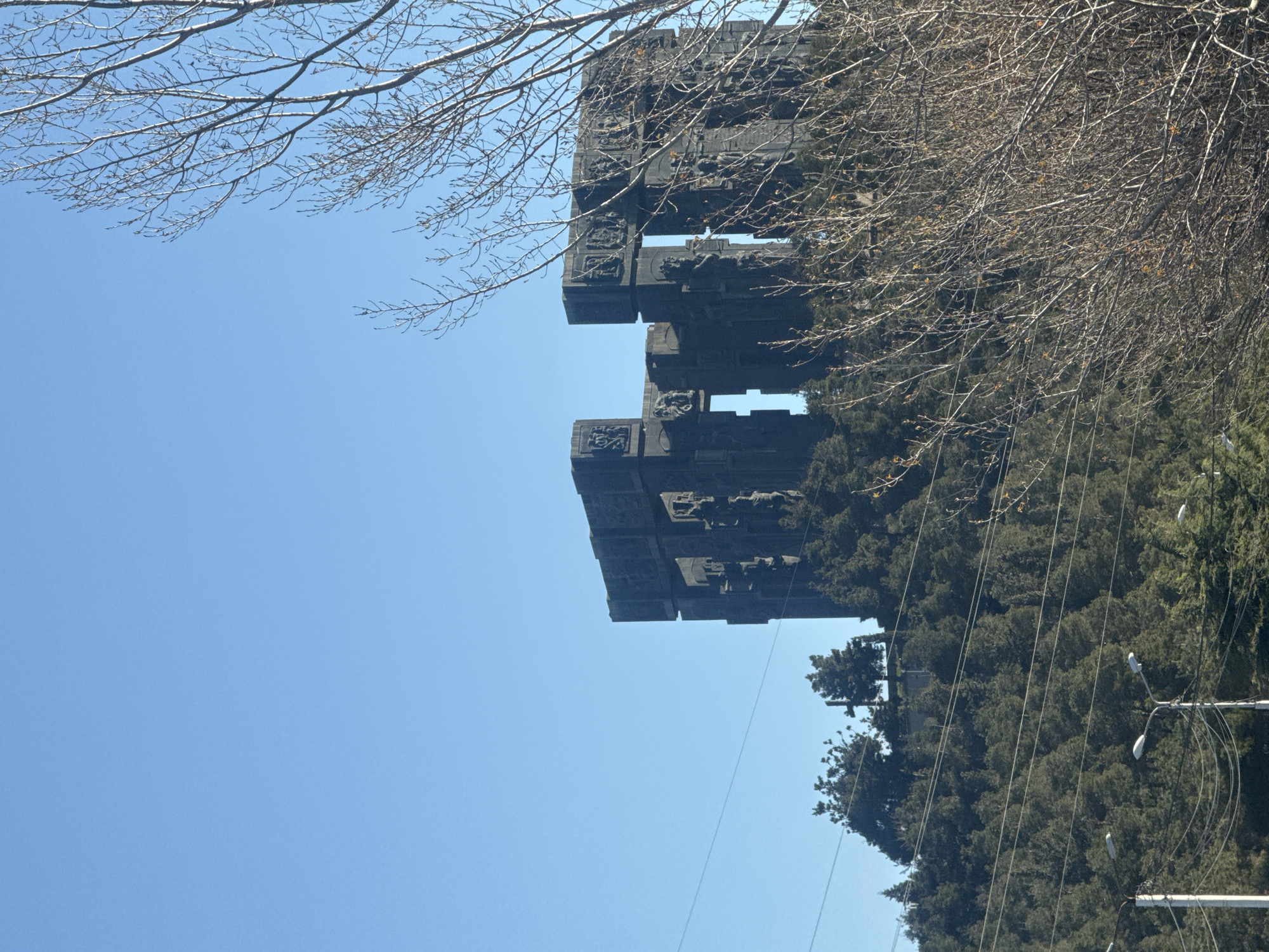 The image shows the Chronicle of Georgia monument, also known as the History Memorial of Georgia or the Stonehenge of Tbilisi, in Tbilisi, Georgia. The monument, composed of large concrete pillars, is set against a clear blue sky and framed by bare tree branches and evergreen trees. Power lines are visible against the background foliage.