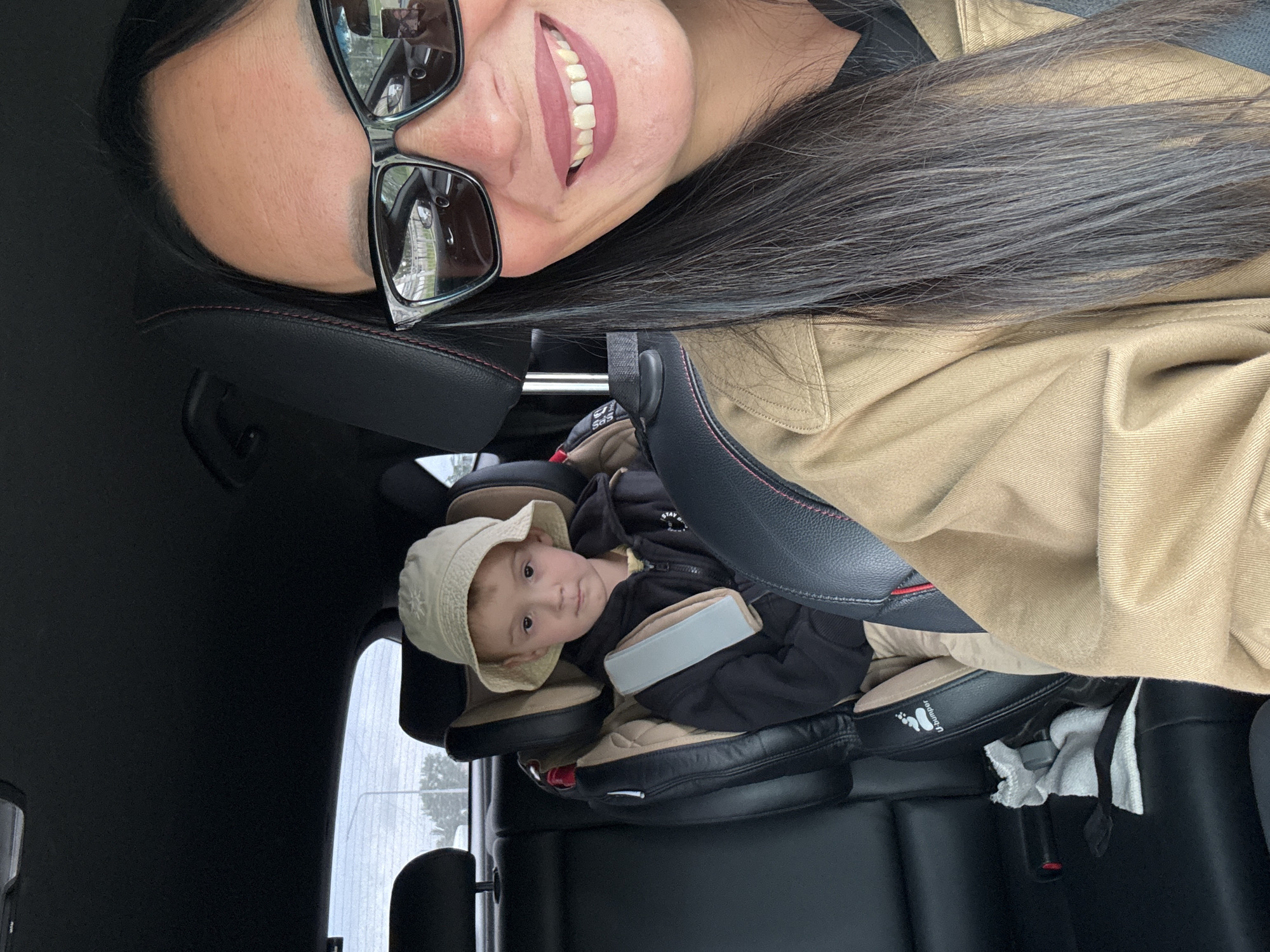 A woman wearing sunglasses and a tan jacket takes a selfie in a car. A young boy wearing a bucket hat and a dark jacket is seated in a car seat behind her. The scene takes place inside a car, suggesting it is likely on a road within or near the city of Chaikovsky, Russia.