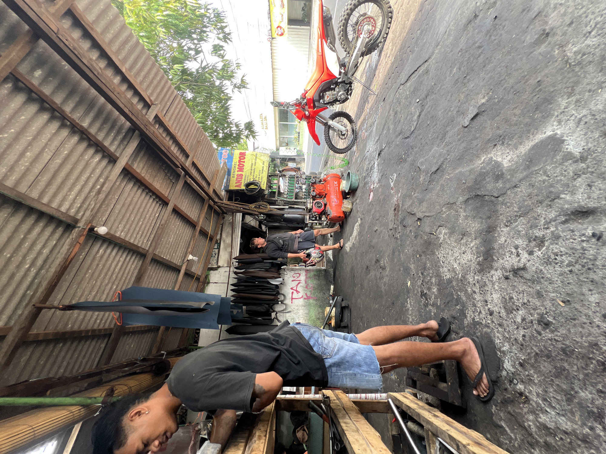 The image depicts the exterior of "BENGKEL ANIS MOTOR" on a street in Denpasar, Indonesia, likely a motorcycle repair shop. The foreground shows a worn, gray asphalt ground covered in dust and debris. To the left, a young man wearing a black t-shirt and denim shorts stands by a wooden workbench filled with tools and equipment. In the midground, another man in a black shirt and dark shorts, holding a red and black helmet, stands on the right. Between them, an orange air compressor and a red dirt bike are parked on the asphalt. The background reveals the shop's covered structure with a corrugated metal roof supported by wooden beams. A sign for "BENGKEL ANIS MOTOR" hangs above the shop, with smaller signs advertising services and products. The scene suggests an active, albeit somewhat cluttered, motorcycle service environment under the bright daylight.